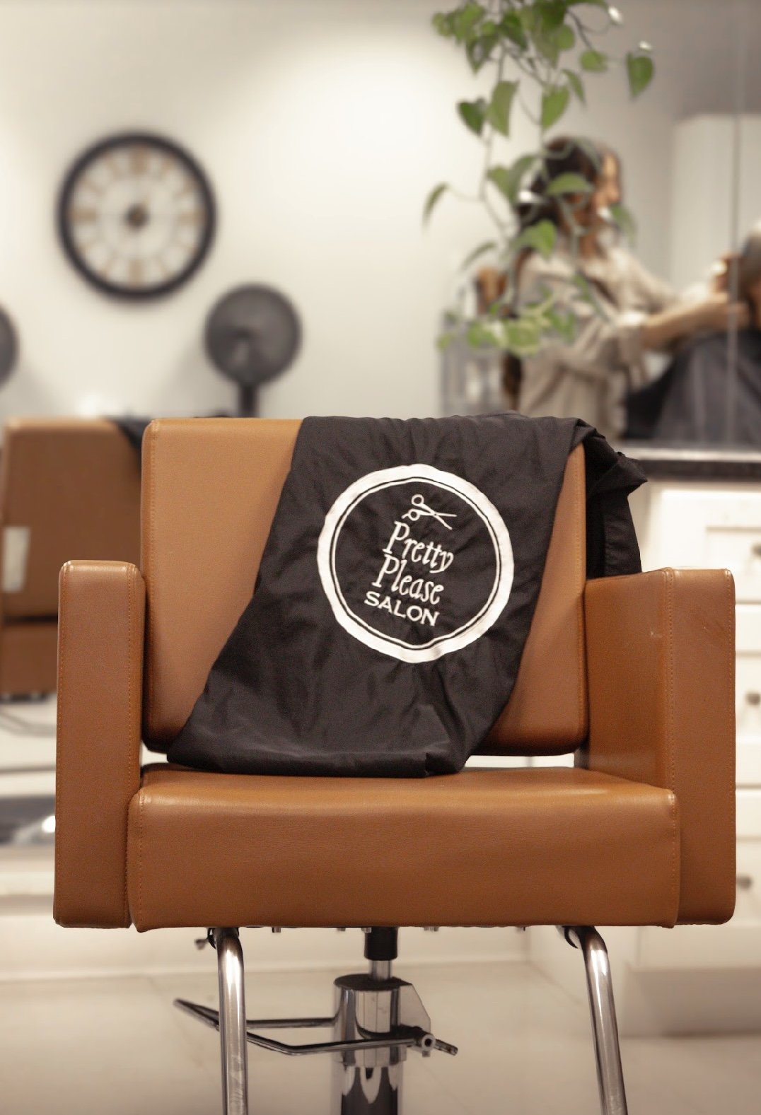 A salon chair with a black cape draped over it, labeled "Pretty Please SALON," in a hair salon with a blurred background including a clock, fan, and a person with leafy plants.
