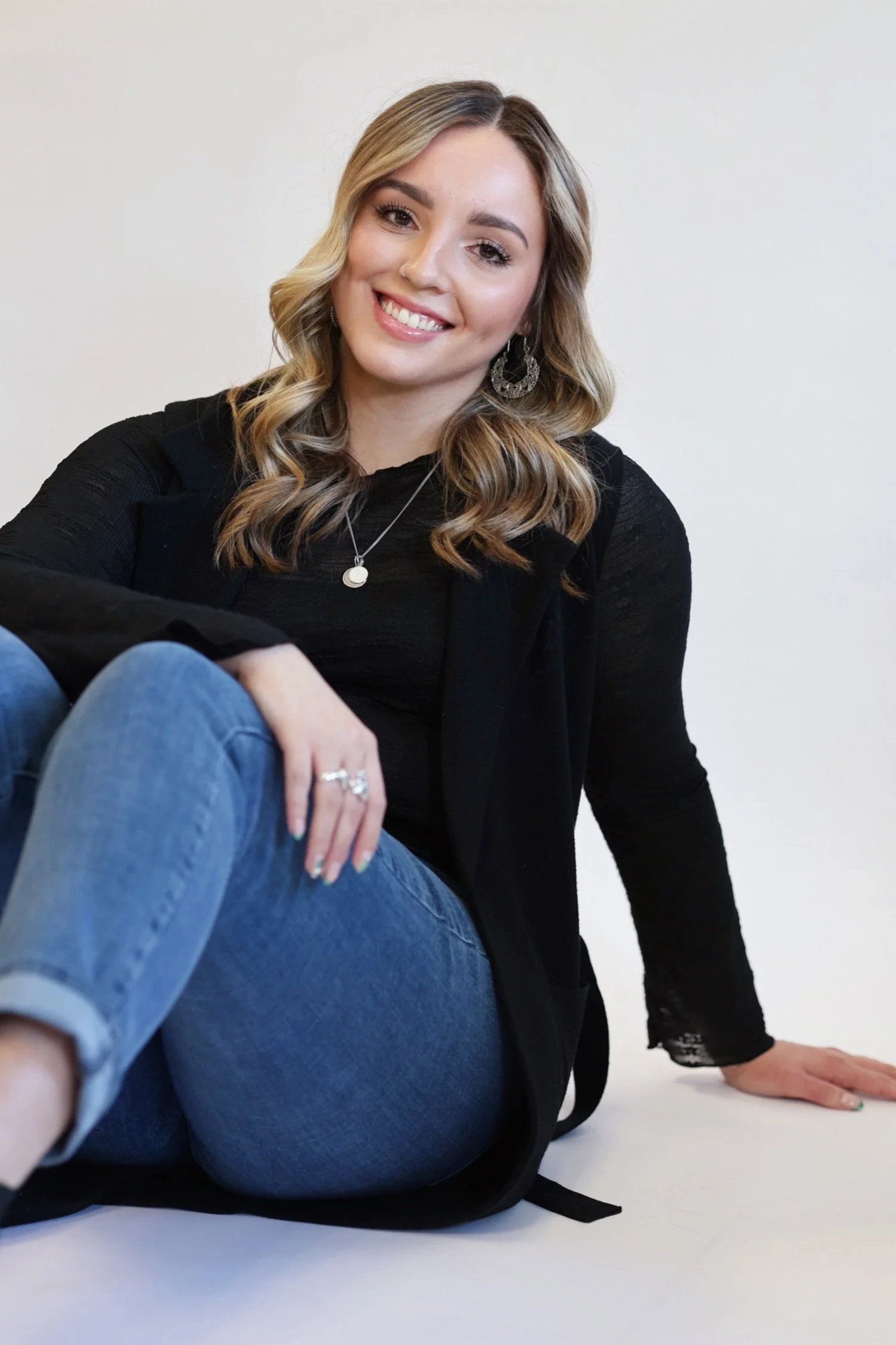 A smiling young woman with long, wavy blonde hair, wearing a black top and silver jewelry, sitting on the floor with one knee up, and a person in jeans lying on the floor in front of her against a plain white background.