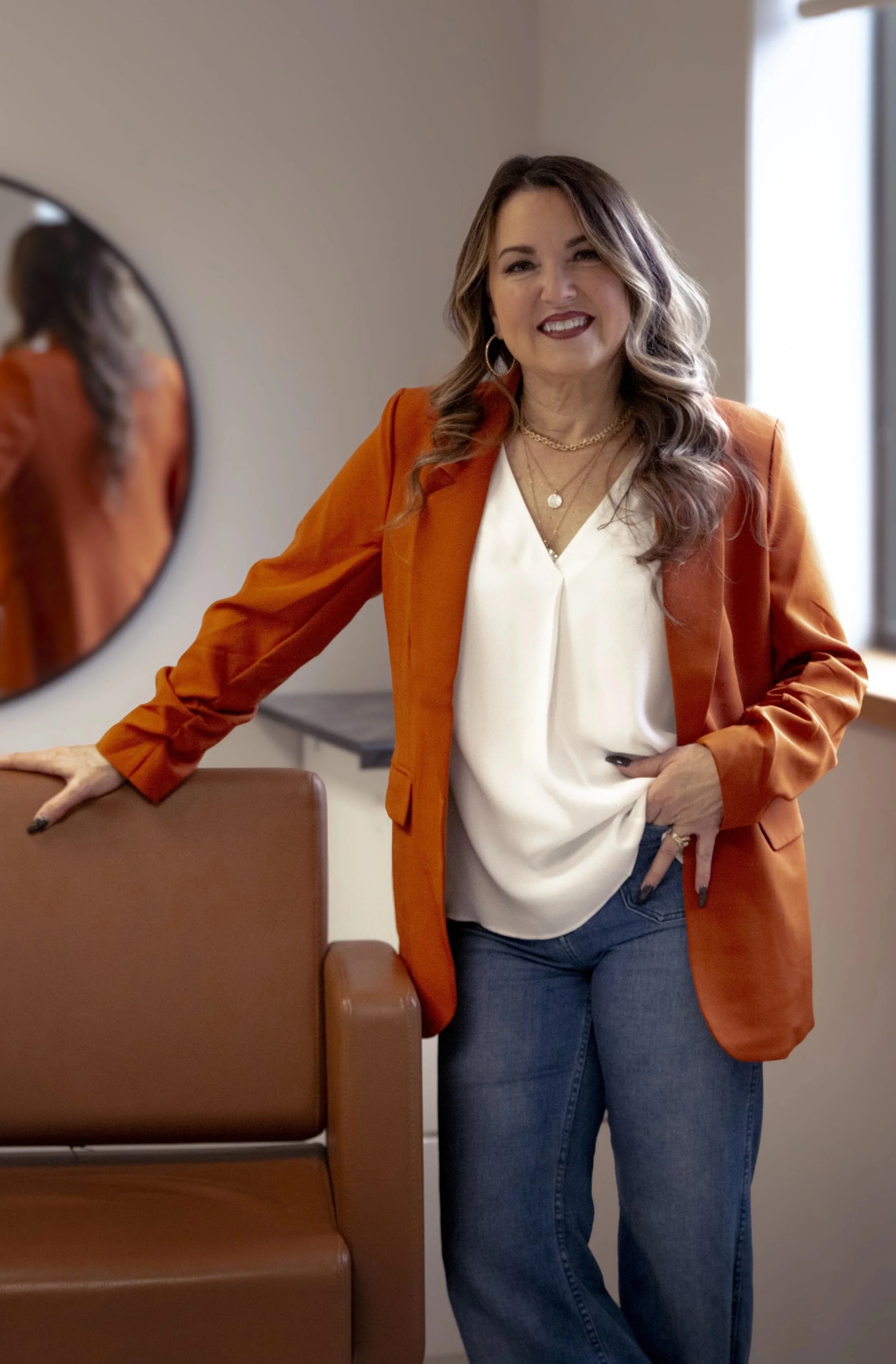 A woman with wavy brown hair, wearing a white blouse and orange blazer, smiling and standing next to a mirror and brown chairs in a room with neutral walls and a window.