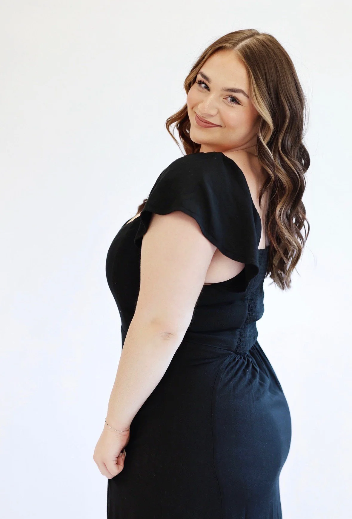 A young woman with long wavy brown hair wearing a black dress with ruffled sleeves, standing against a plain white background, looking over her shoulder and smiling.