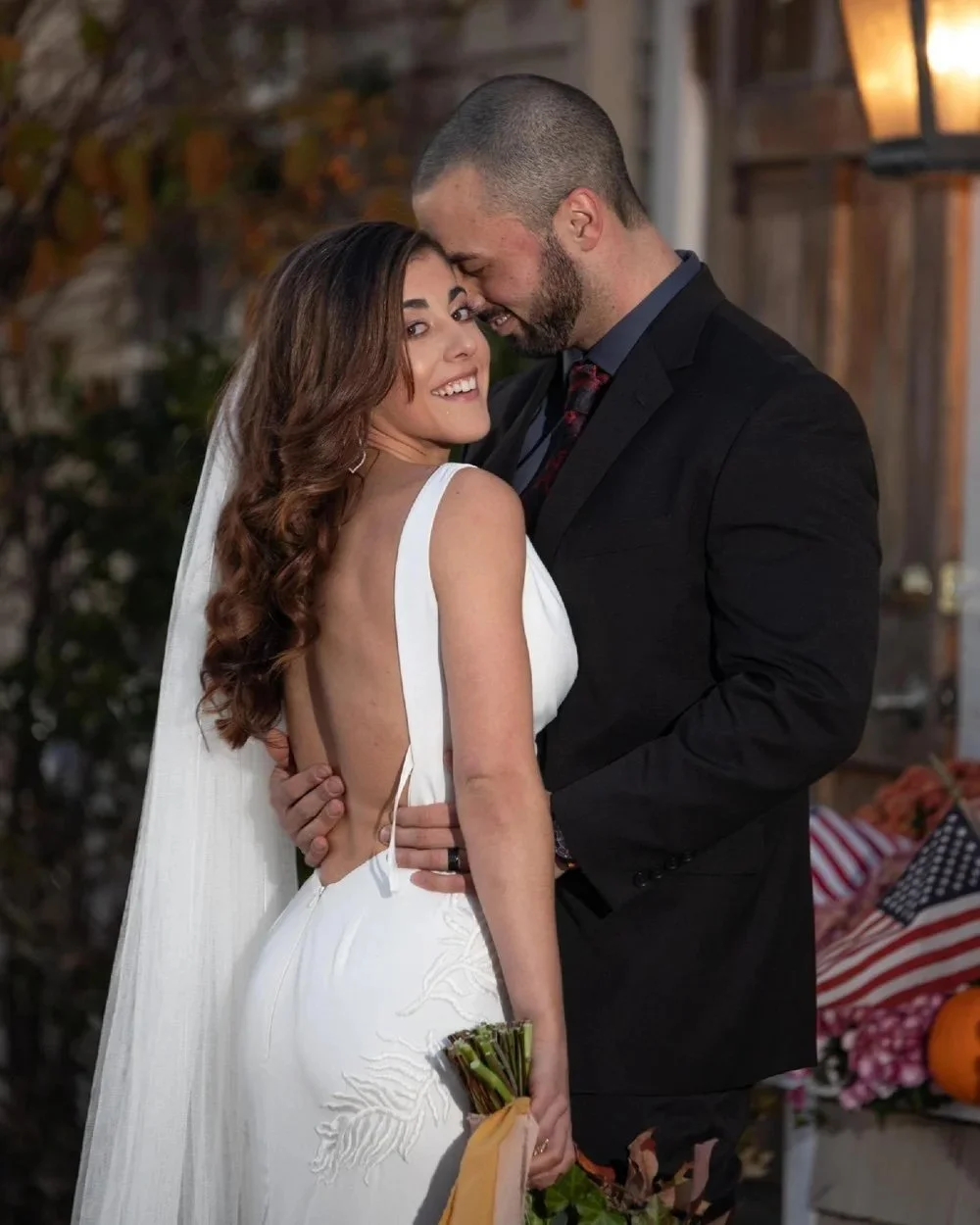 A bride and groom sharing a romantic moment at their wedding, with the bride smiling and holding a bouquet of flowers, and the groom leaning in with their foreheads touching, in an outdoor setting.