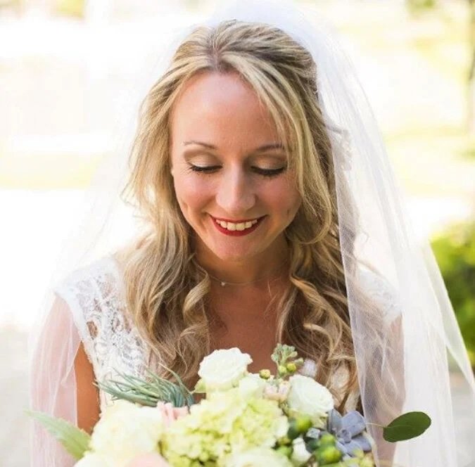 A smiling bride with blonde hair and a lace wedding dress holding a bouquet of white and light pink flowers in an outdoor setting.
