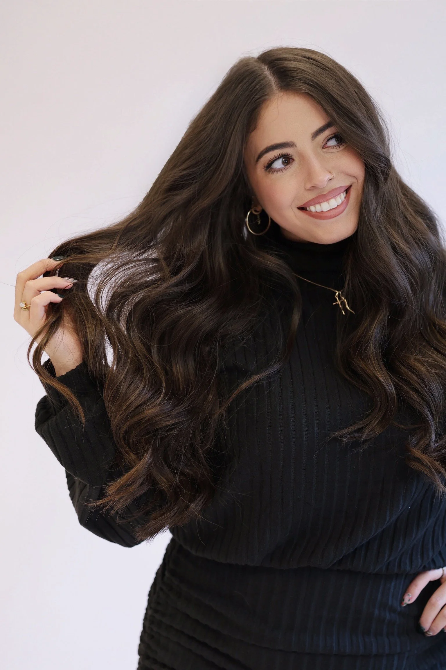 A young woman with long, wavy brown hair, wearing a black turtleneck sweater, hoop earrings, and a gold necklace, is smiling and holding her hair with one hand.