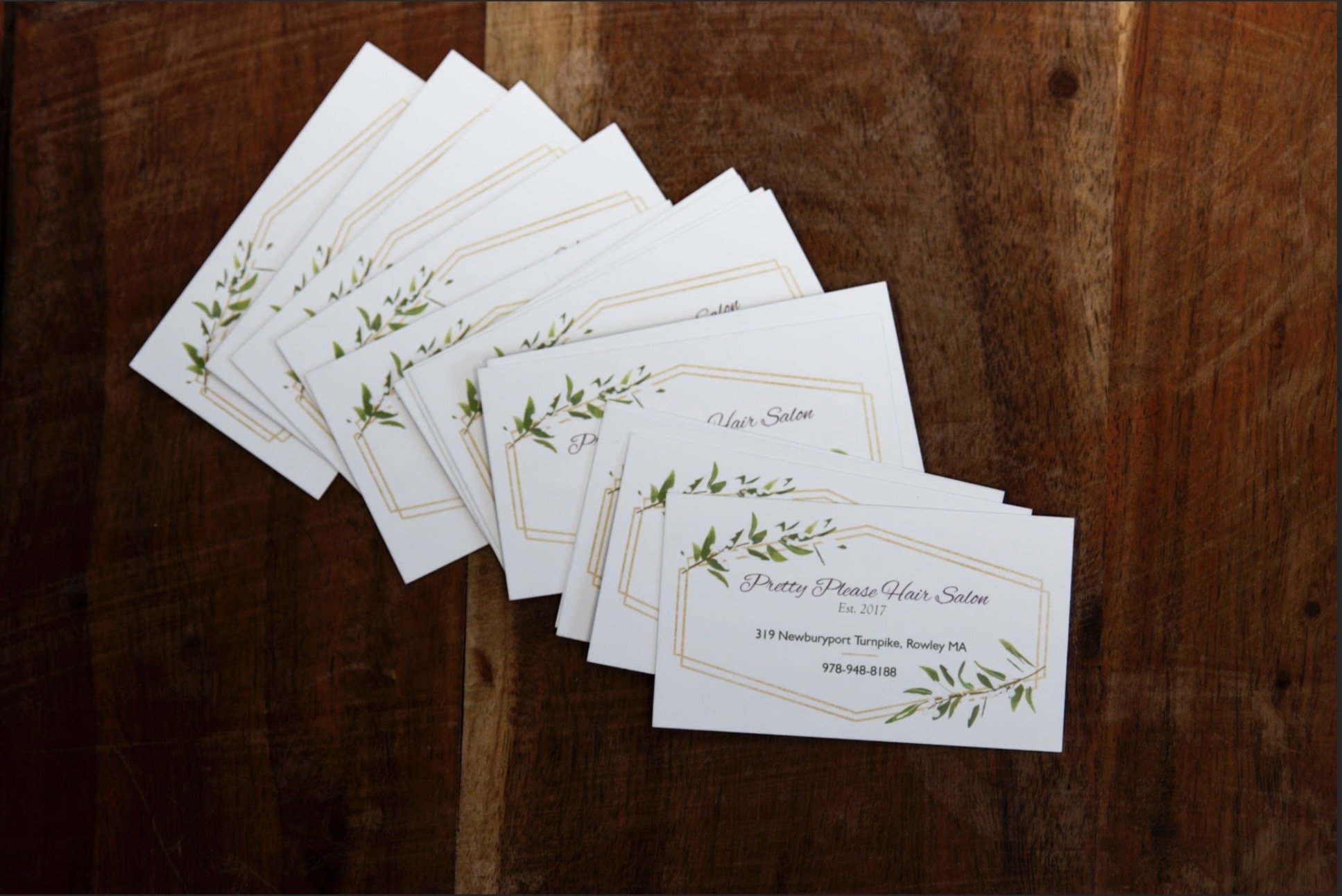 A stack of printed business cards placed on a wooden surface. The cards are for Pretty Please Hair Salon, including an address, phone number, and decorative green leaves.