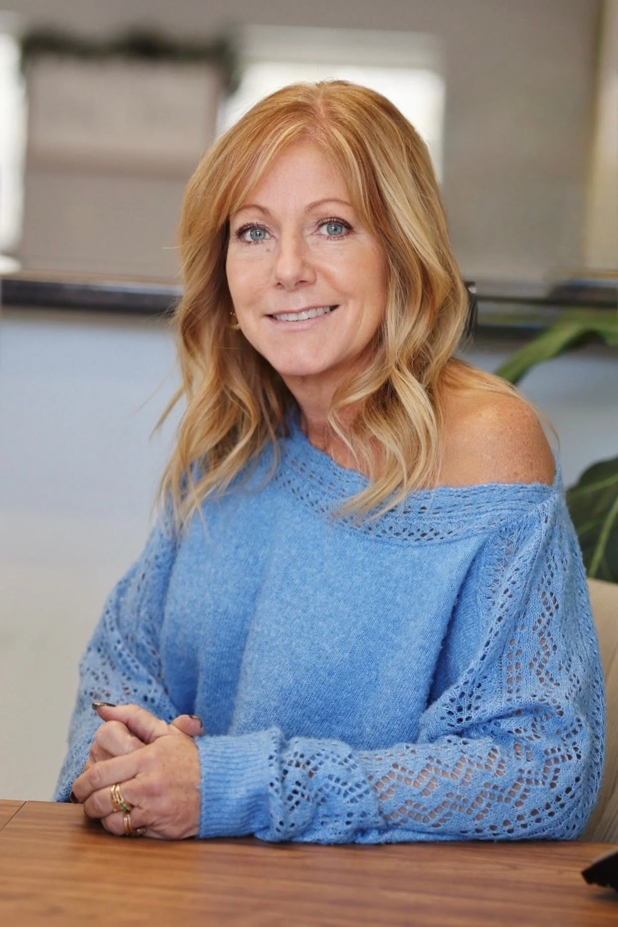 A smiling middle-aged woman with blonde hair and blue eyes, wearing a blue open-knit sweater, sitting at a wooden table.