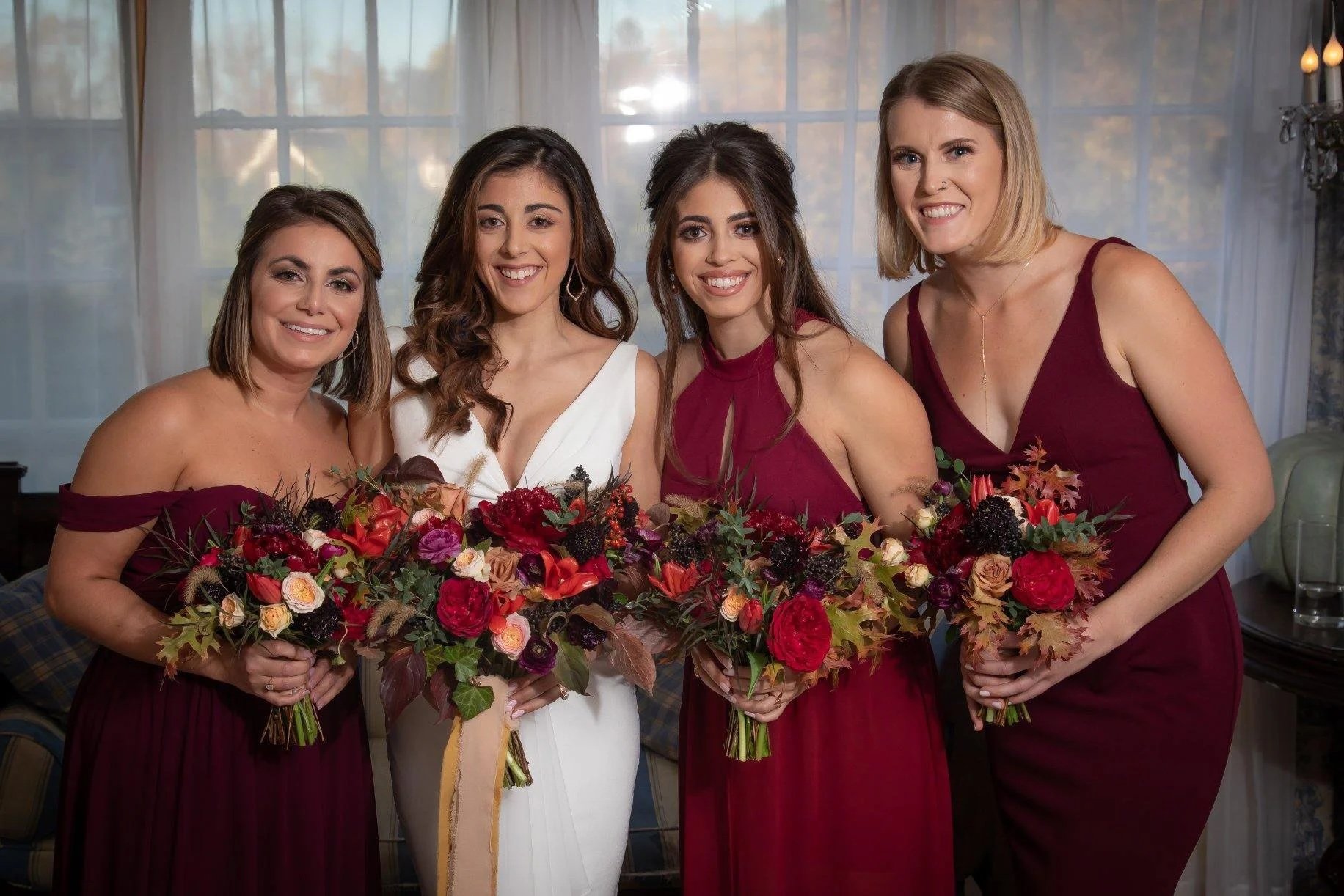 Four women dressed in formal attire, holding large bouquets of autumn-colored flowers, standing in a room with large windows and white curtains.