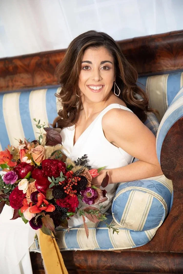 A woman with brown wavy hair and a white dress sitting on a blue and beige upholstered couch, holding a colorful bouquet of flowers, smiling at the camera.