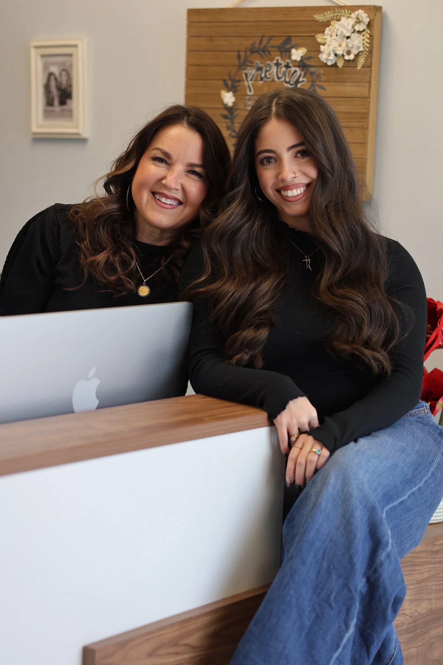 Two women smiling in front of a decorative wooden wall hanging with the word 'pretend' and white flowers, sitting at a table with a laptop.