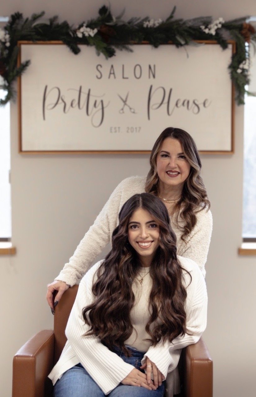 Two women smiling in an indoor setting with a sign that reads 'Salon Pretty Please' in the background, decorated with greenery and white flowers.