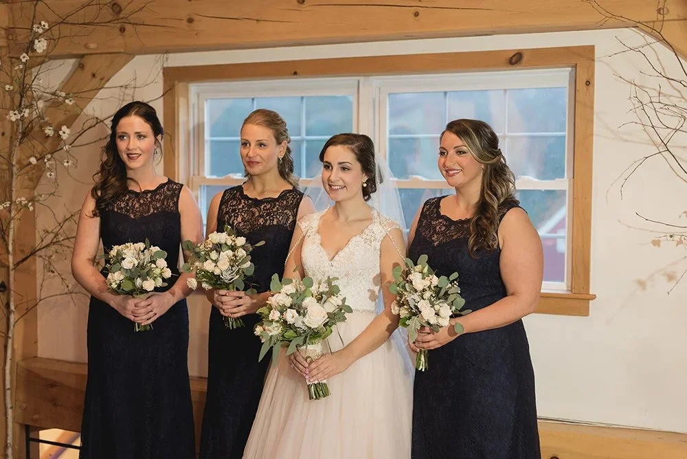 A bride and three bridesmaids standing indoors, holding bouquets, with wooden walls and a window in the background.