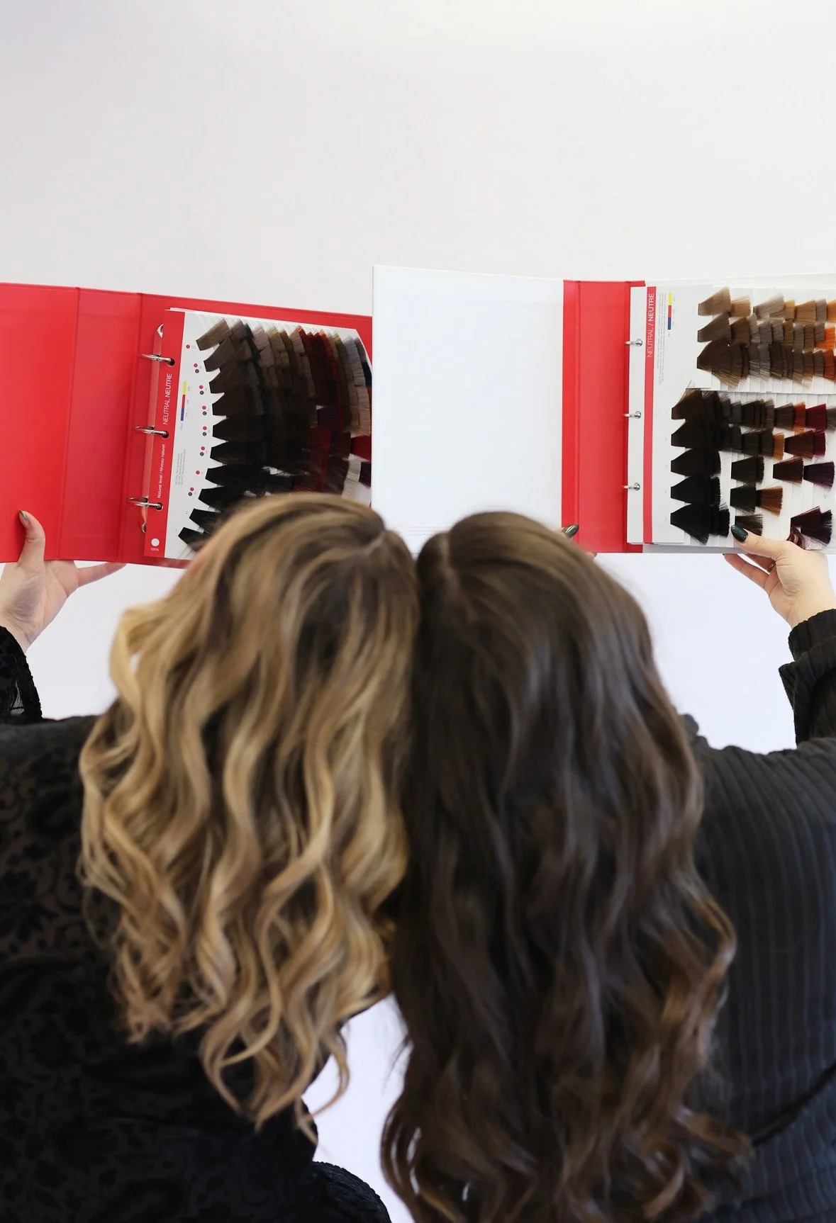 Two women with wavy hair looking at a color hair swatch book while holding it open.