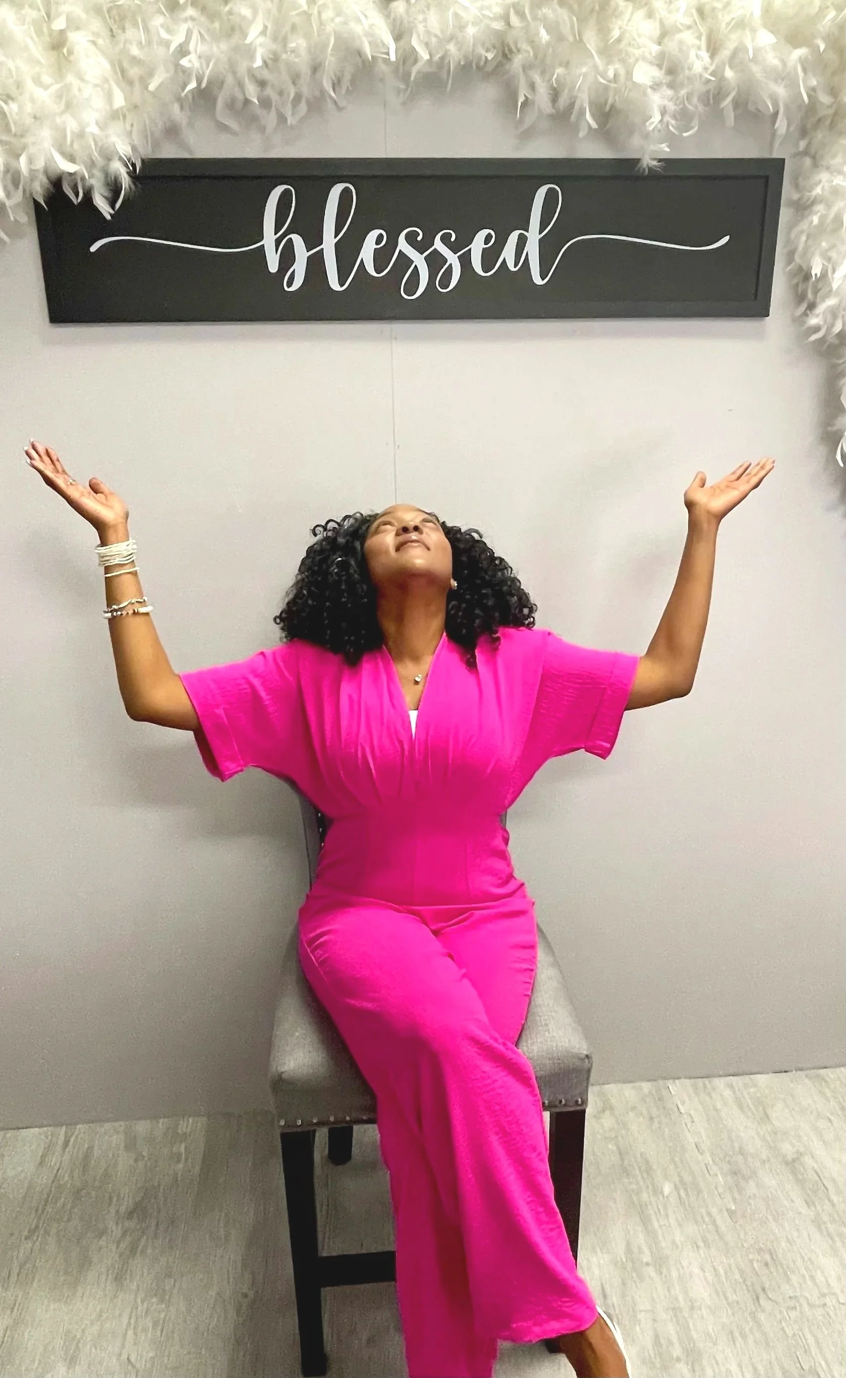 Sheenayvonne sitting in a chair with her hands up towards the black and white "blessed" sign on the wall behind her.