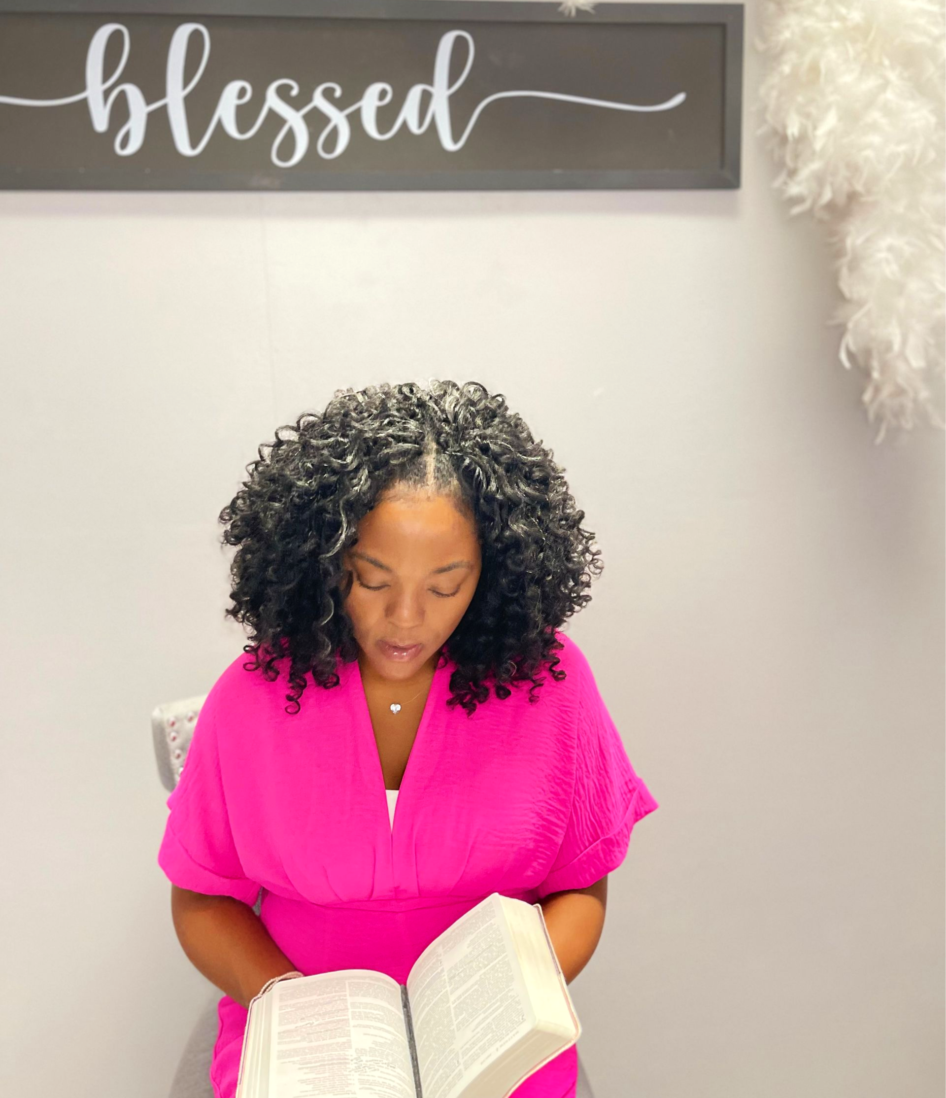 Sheenayvonne sitting in a chair, reading the bible with the black and white "blessed" sign on the wall behind her.