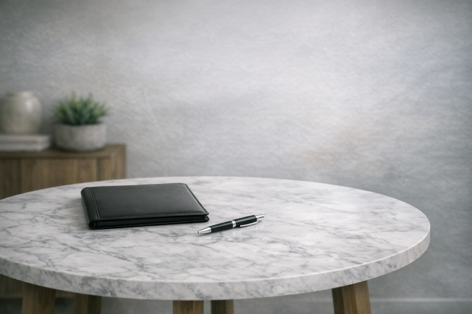 marble top table with a black portfolio and pen laying on desk. Also showing a wood console with a green plant in a marble pot.