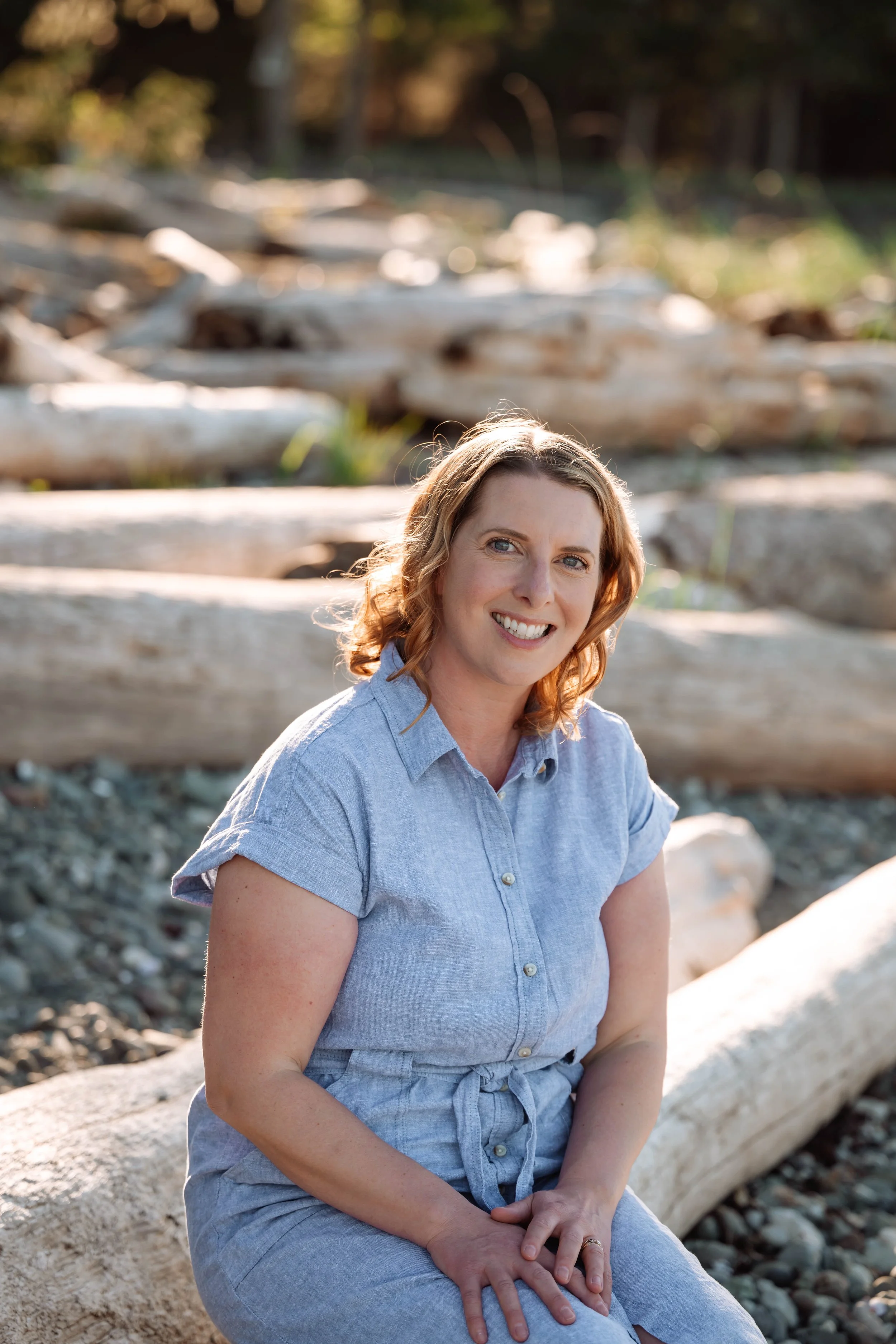 Alyson smiling, sitting on driftwood at the beach in a blue shirt