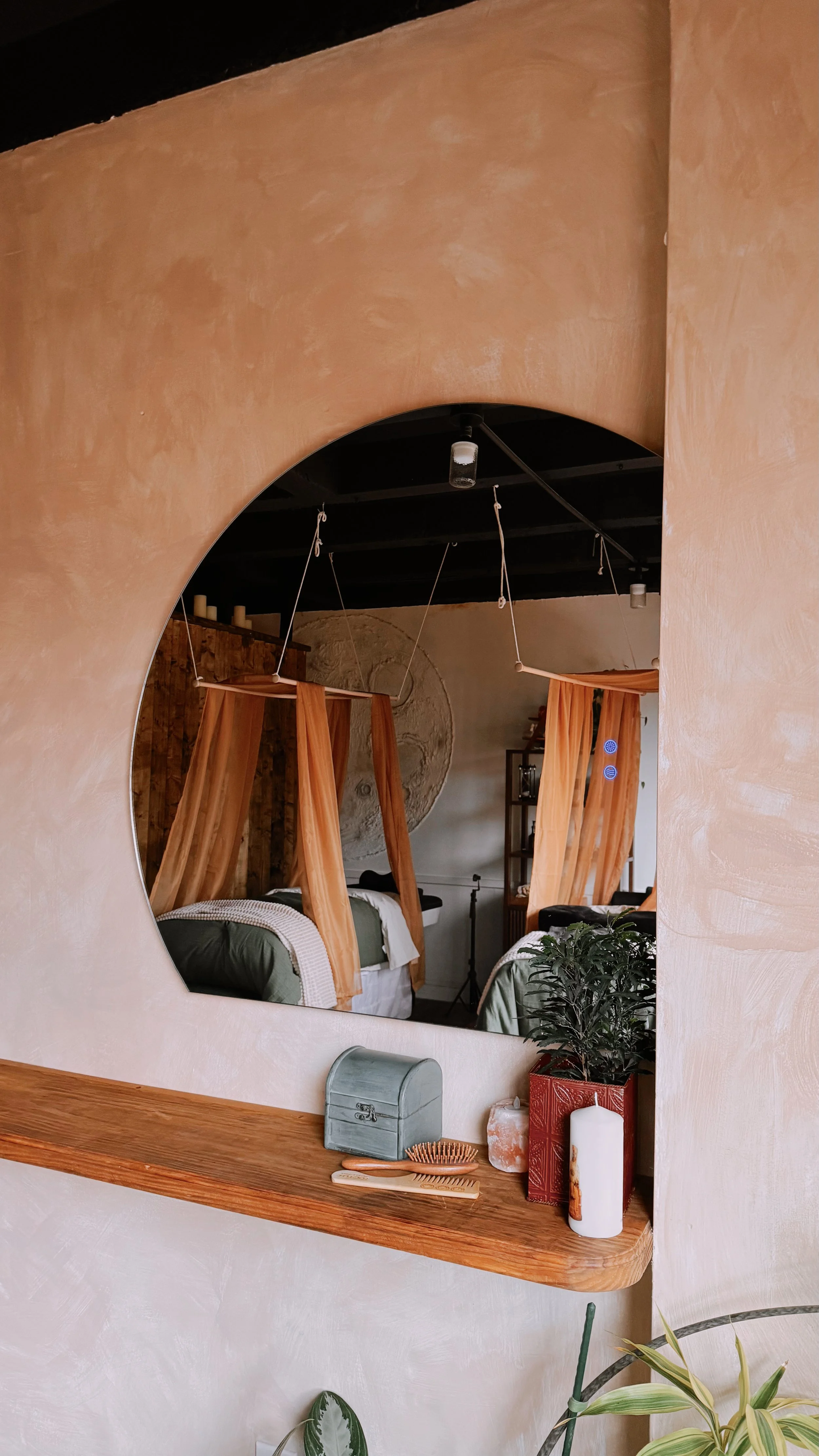 View through a circular cutout in a wall showing a bedroom with two beds with orange curtains, a large circular wall art, and a shelf with plants and decor in the background.