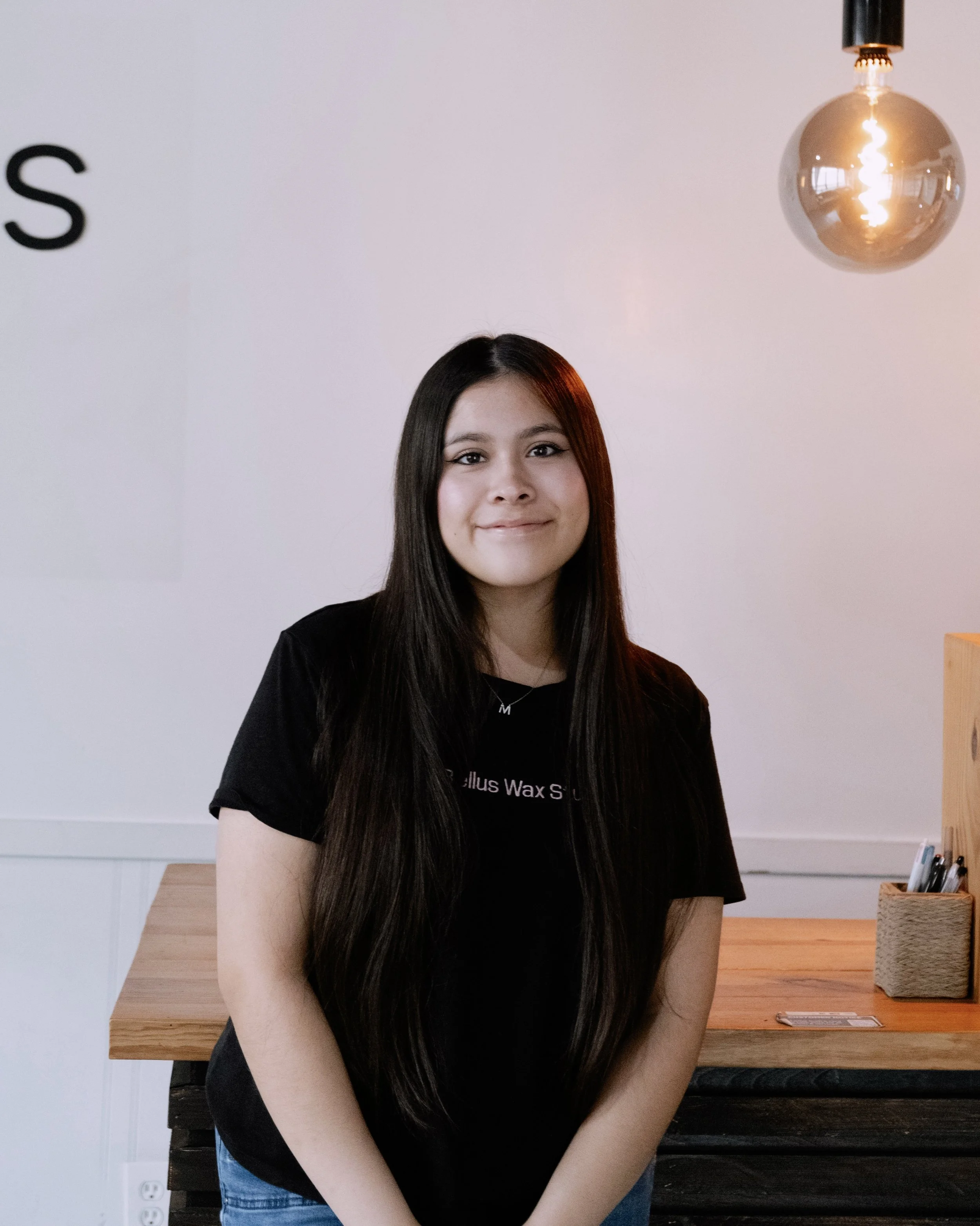 A young woman with long dark hair standing indoors, smiling at the camera. She is wearing a black t-shirt and is in a room with a white wall, wooden furniture, and a modern hanging light fixture.