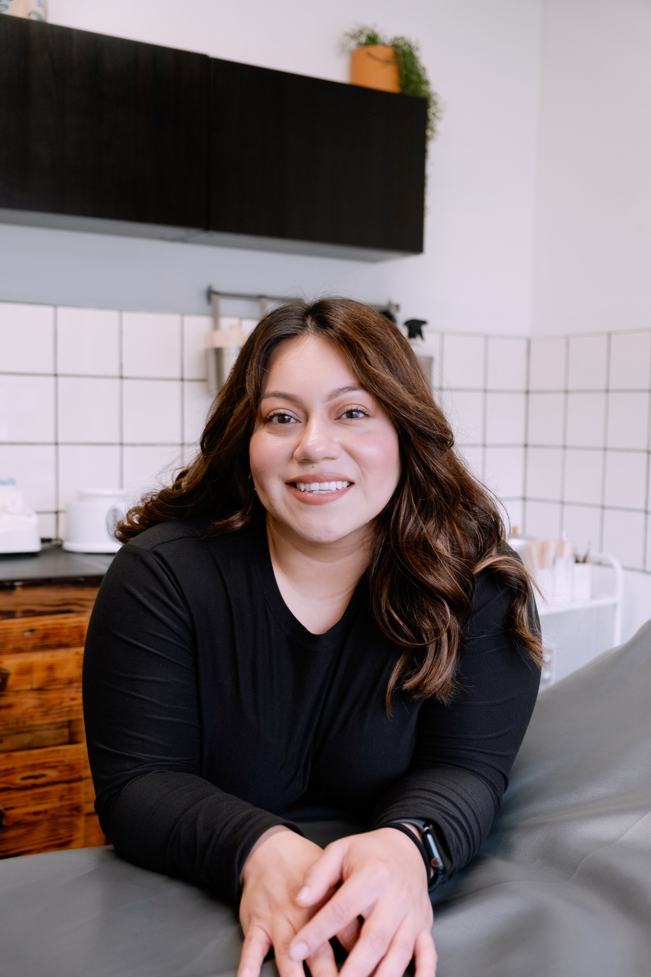 A woman with brown, wavy hair smiling and sitting on a gray couch in a modern kitchen setting with white tile walls and dark cabinets.