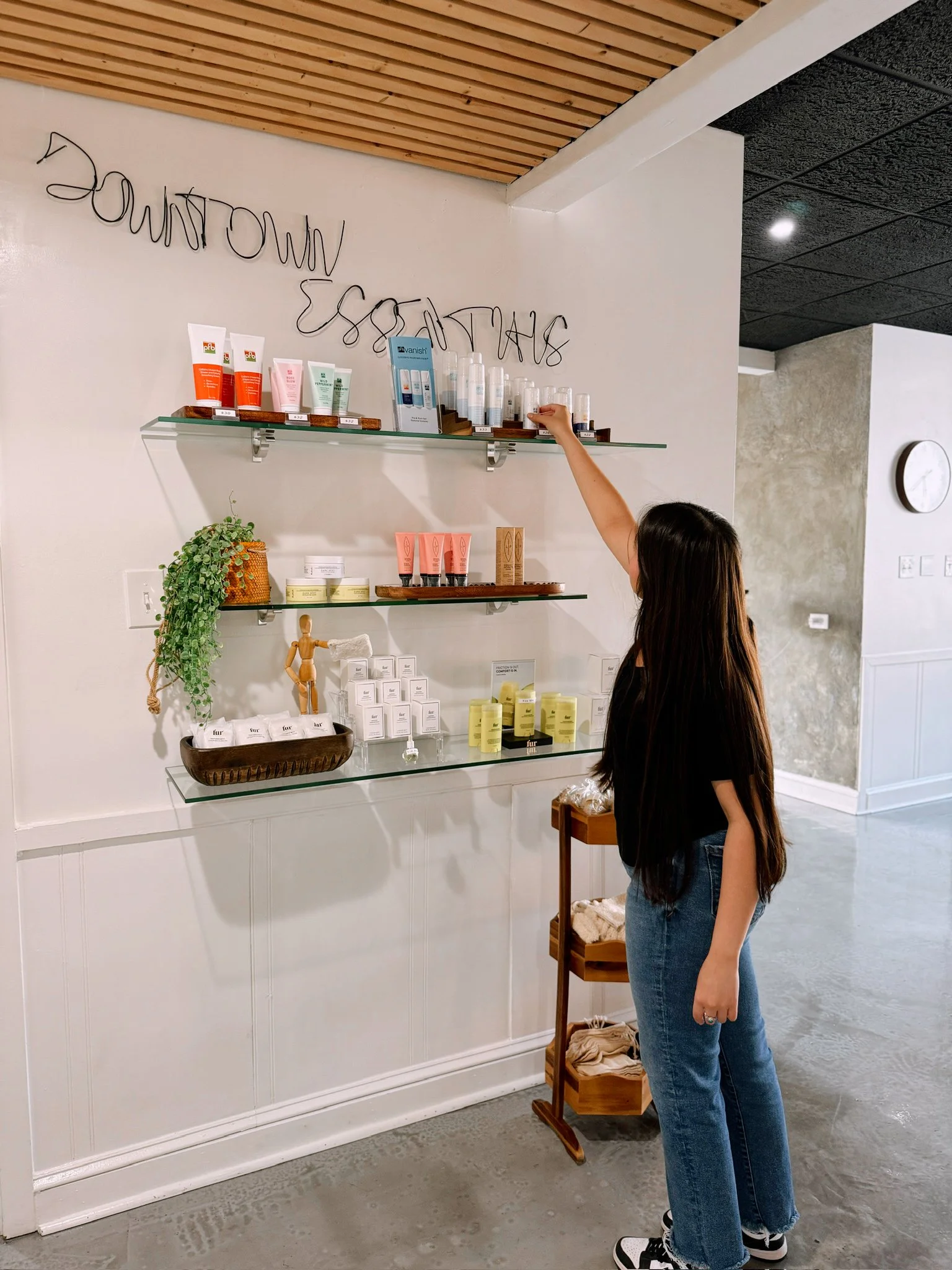 A woman with long dark hair wearing a black T-shirt and jeans browsing skincare products on glass shelves in a spa or beauty store, along with a small wooden cart with towels.