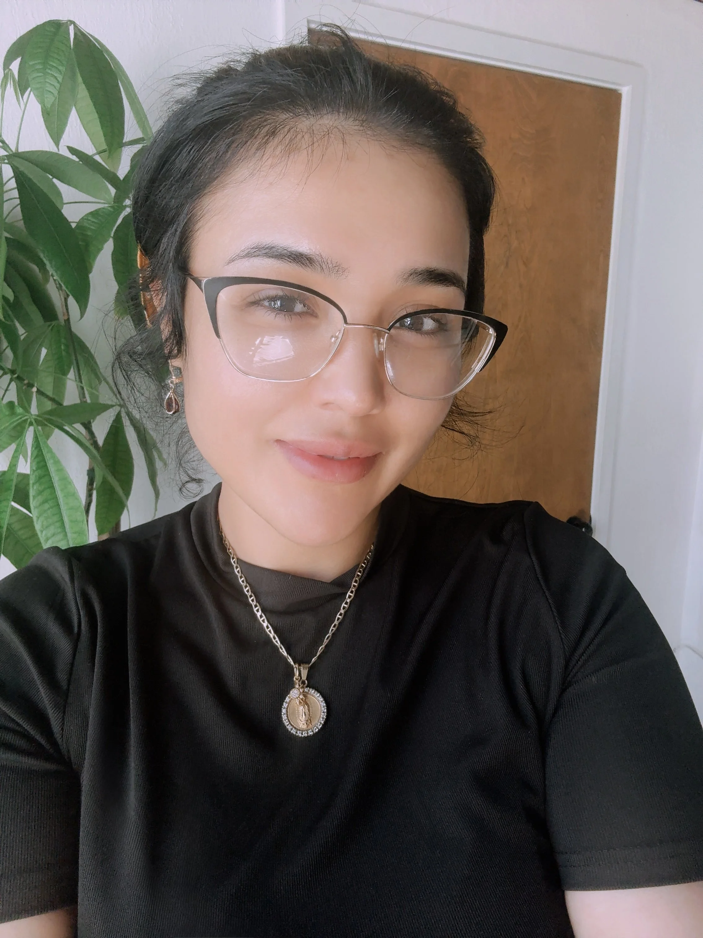 Close-up of a young woman with glasses, wearing a black shirt, gold jewelry, standing indoors near a plant with green leaves and a wooden door in the background.