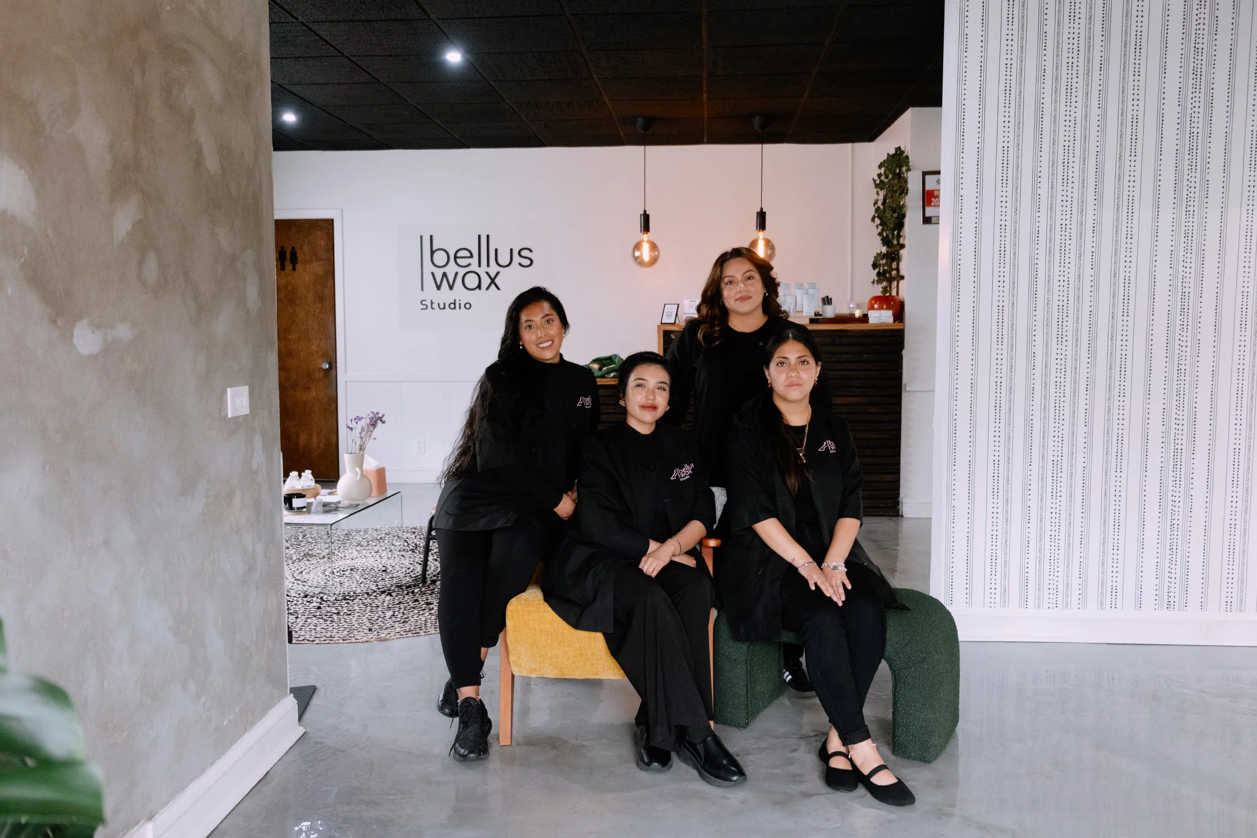 Four women in black uniforms sitting and standing in a modern salon or studio with a sign that reads "bellus wax Studio" in the background.