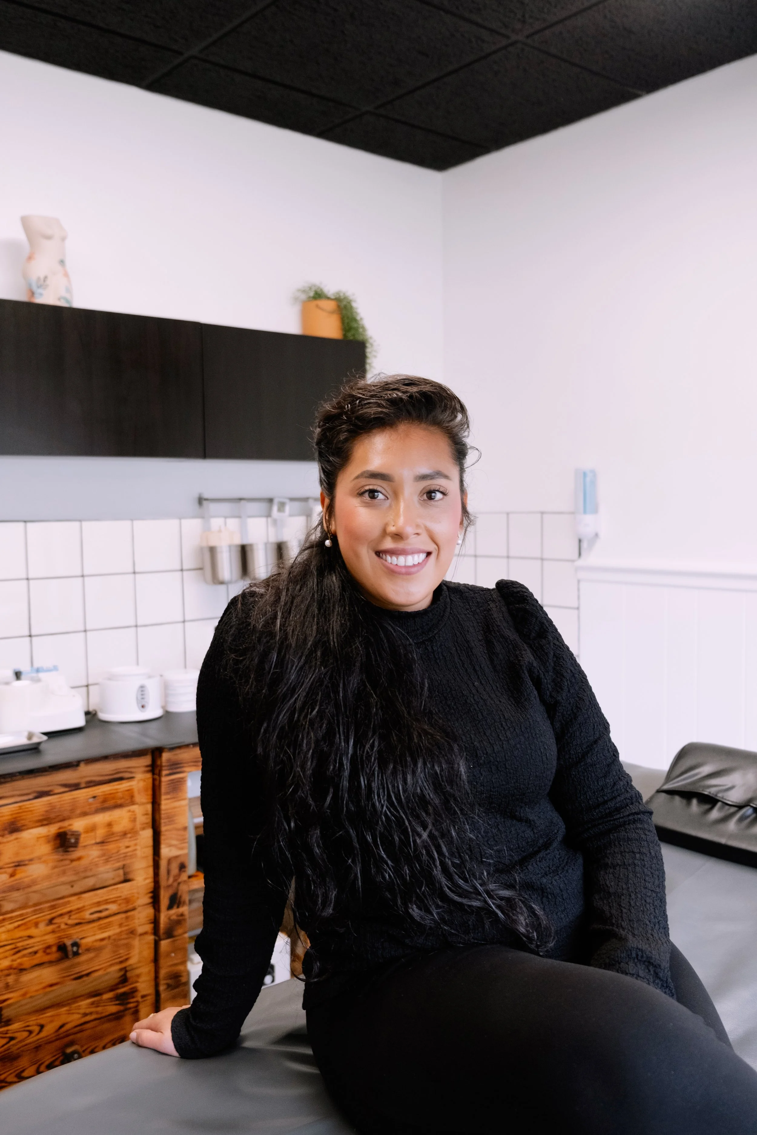 A woman with long dark hair, wearing a black sweater, is sitting on a medical examination table, smiling in a healthcare or medical office.
