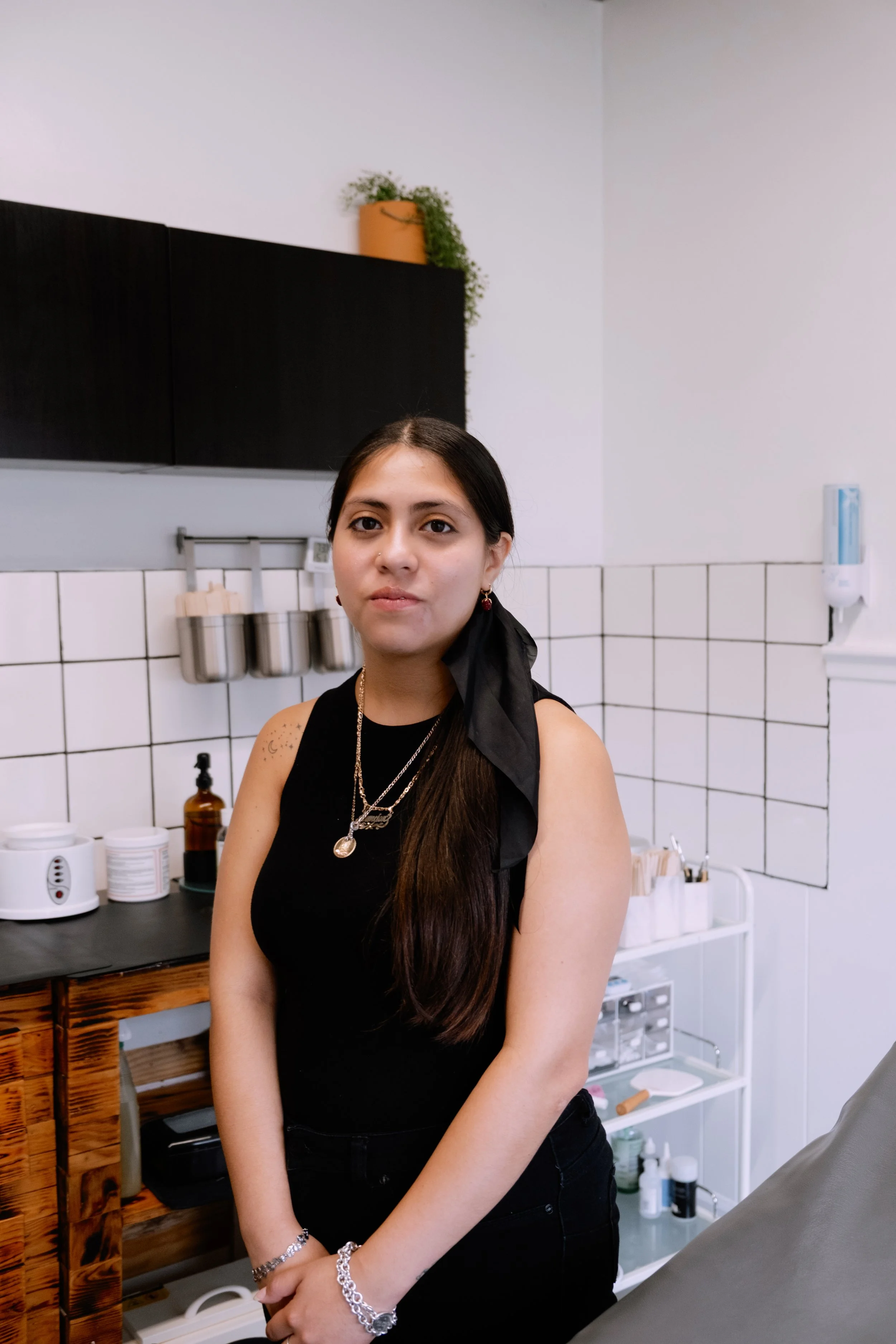 A young woman with long dark hair wearing a black sleeveless top and jewelry, standing in a medical or spa clinic with white tiled walls and medical supplies in the background.