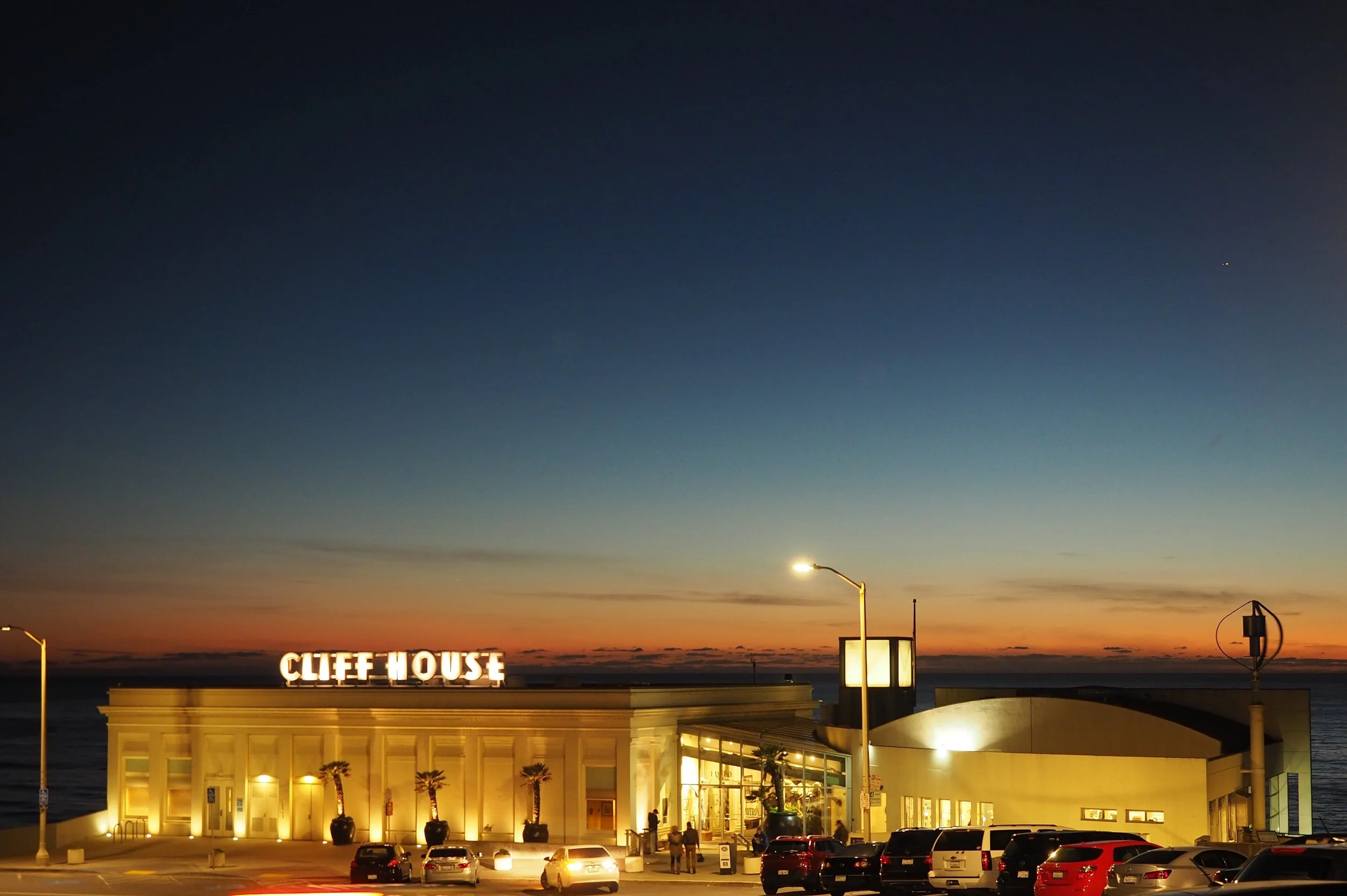 Sunset over a building with a sign that reads 'CLIFF HOUSE', overlooking a parking lot with cars and streetlights, with a body of water in the background.