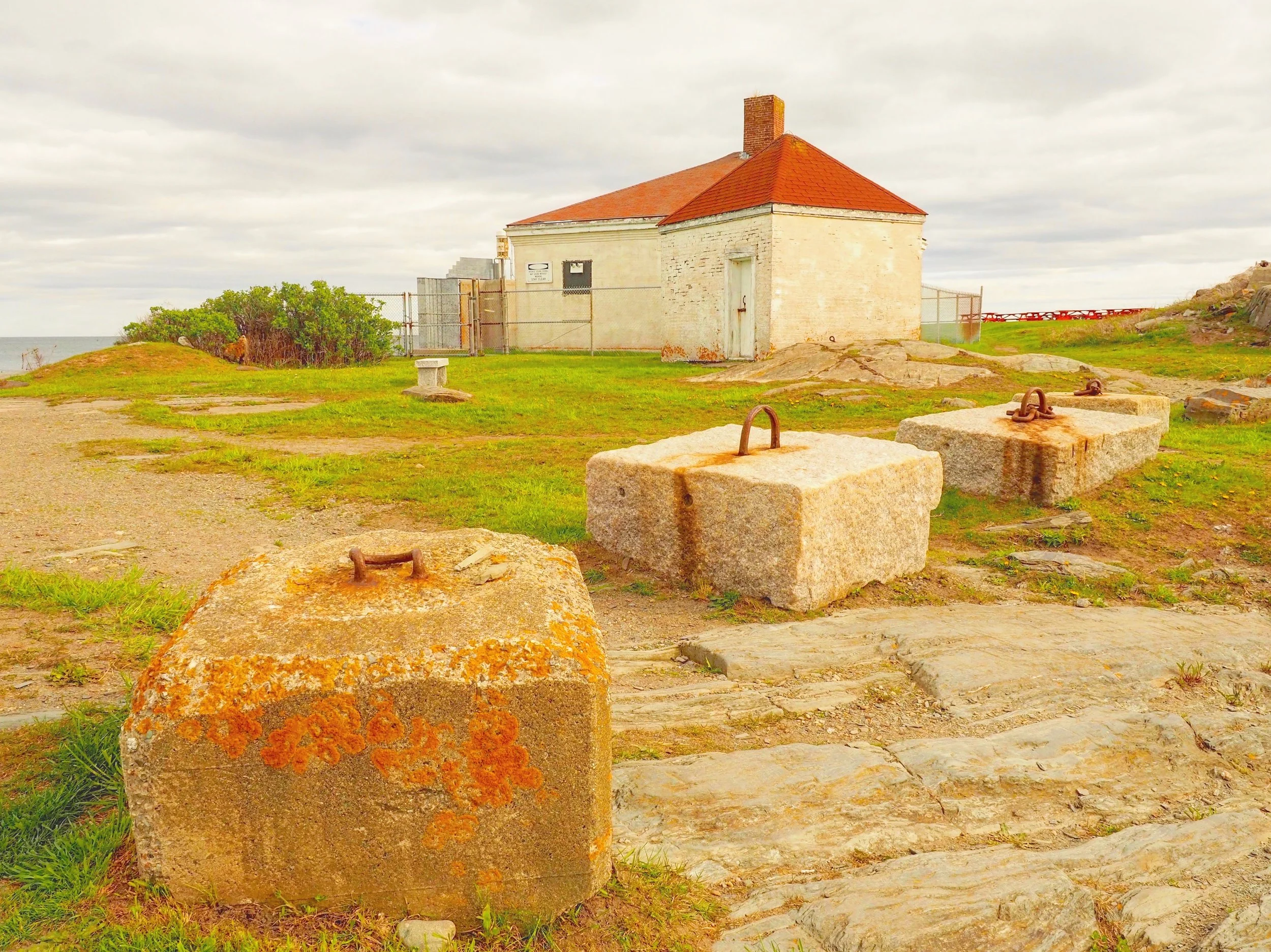 A small white brick building with a red sloped roof and a brick chimney, surrounded by a wire fence, with green grass, rocks, and a cloudy sky in the background.
