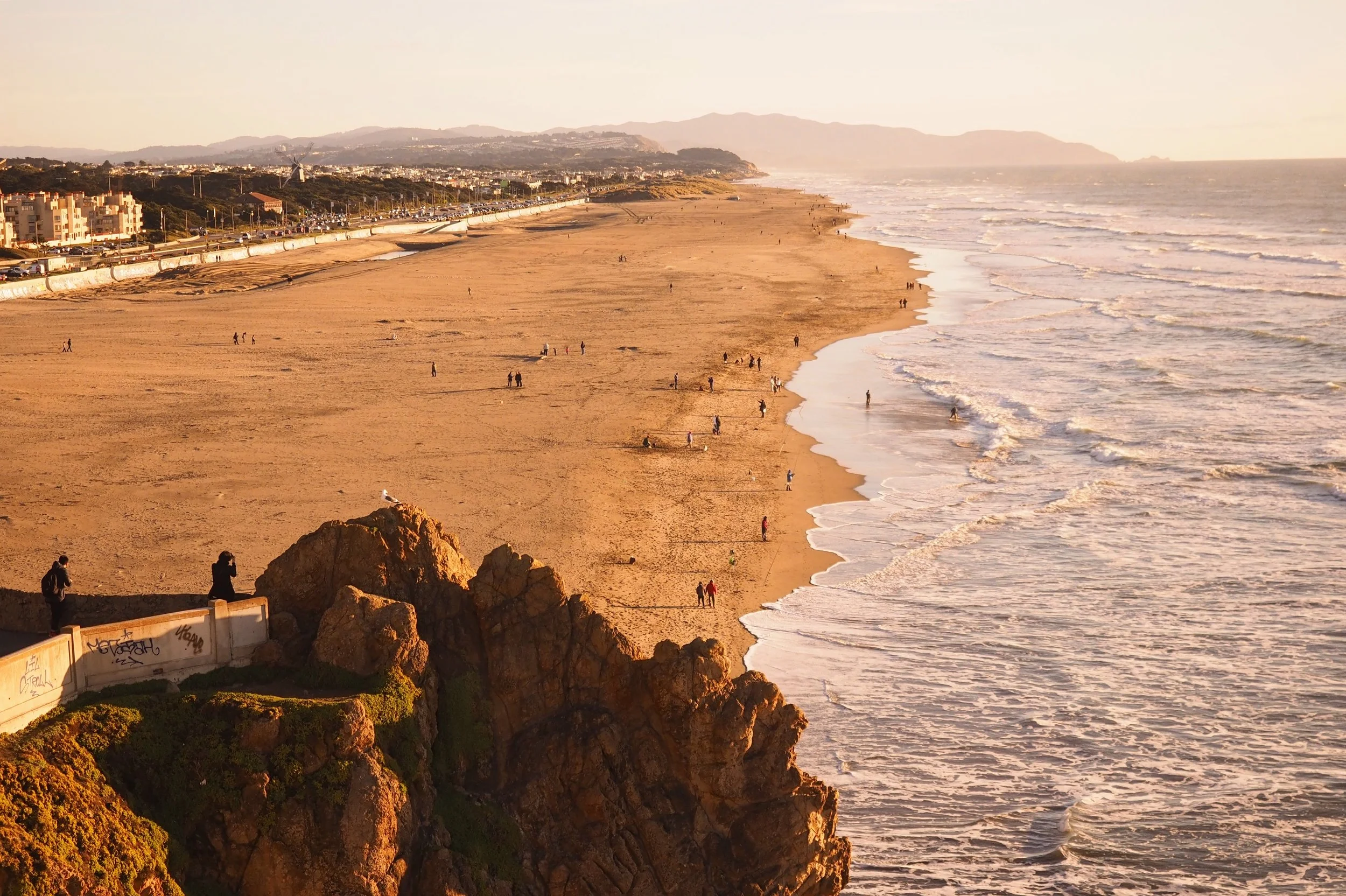 A scenic view of a beach at sunset with people walking along the shoreline and a rocky cliff in the foreground.