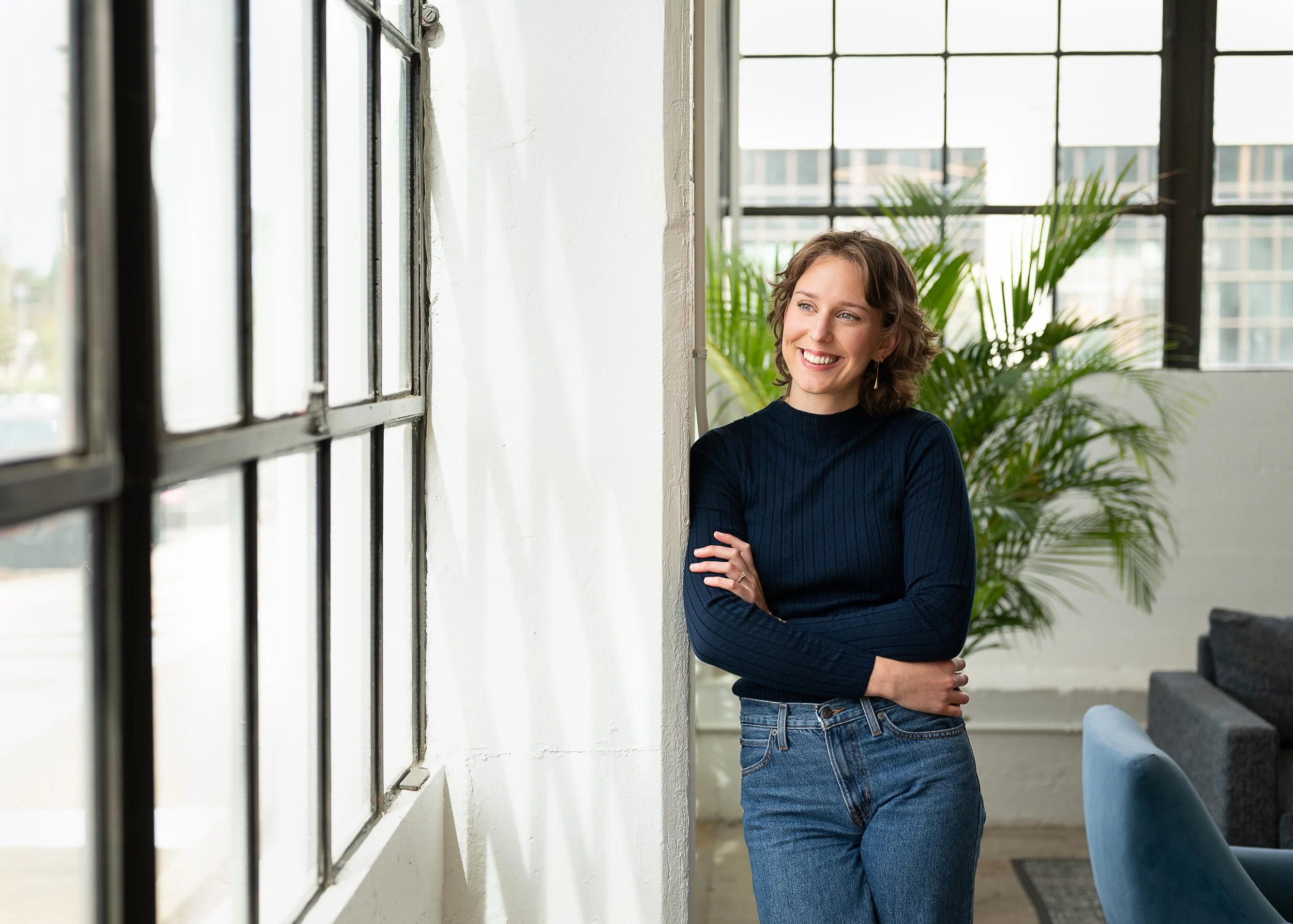 A woman with short curly hair wearing a dark blue sweater and jeans, smiling and looking out of a large industrial-style window in a bright, modern room with green plants.