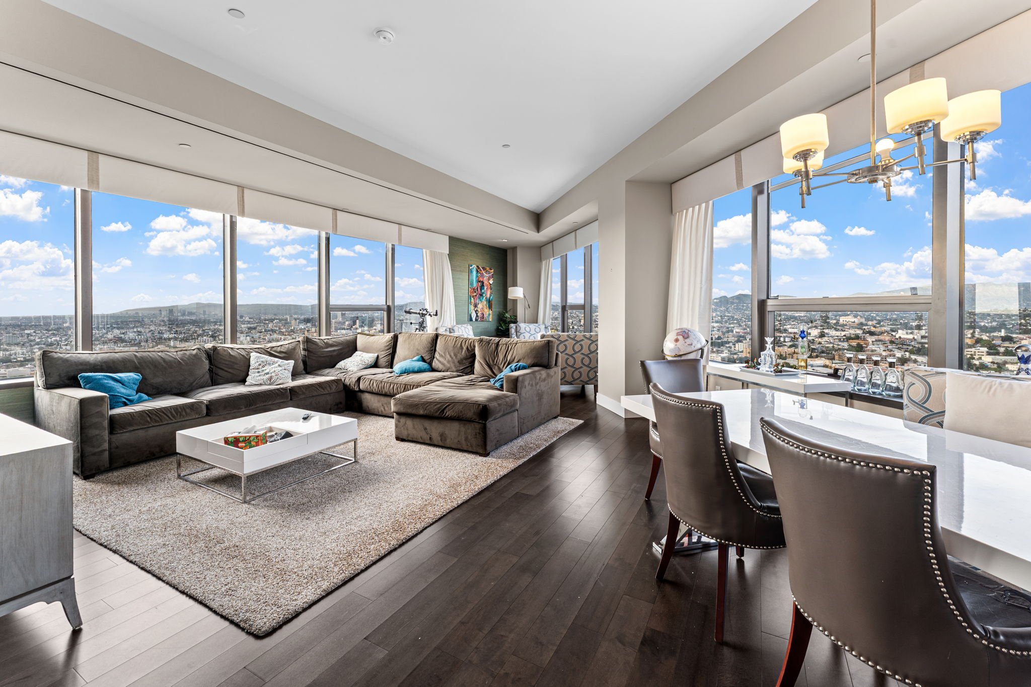Modern high-rise apartment living room with large windows, brown sectional sofa, white coffee table, area rug, and dining table with chairs, cityscape view outside.