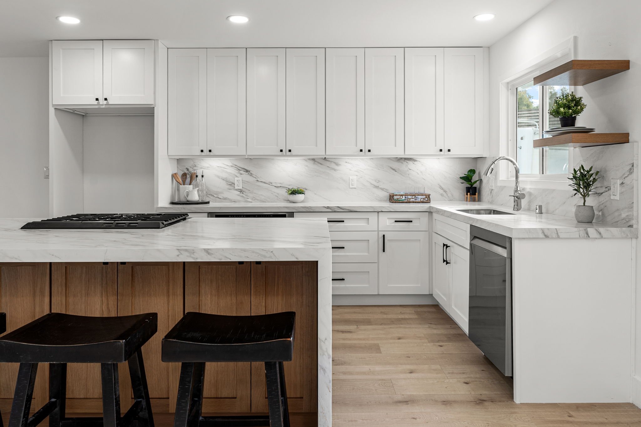 Modern kitchen with white cabinets, marble backsplash and countertops, wooden island with black stools, and a window with plants.