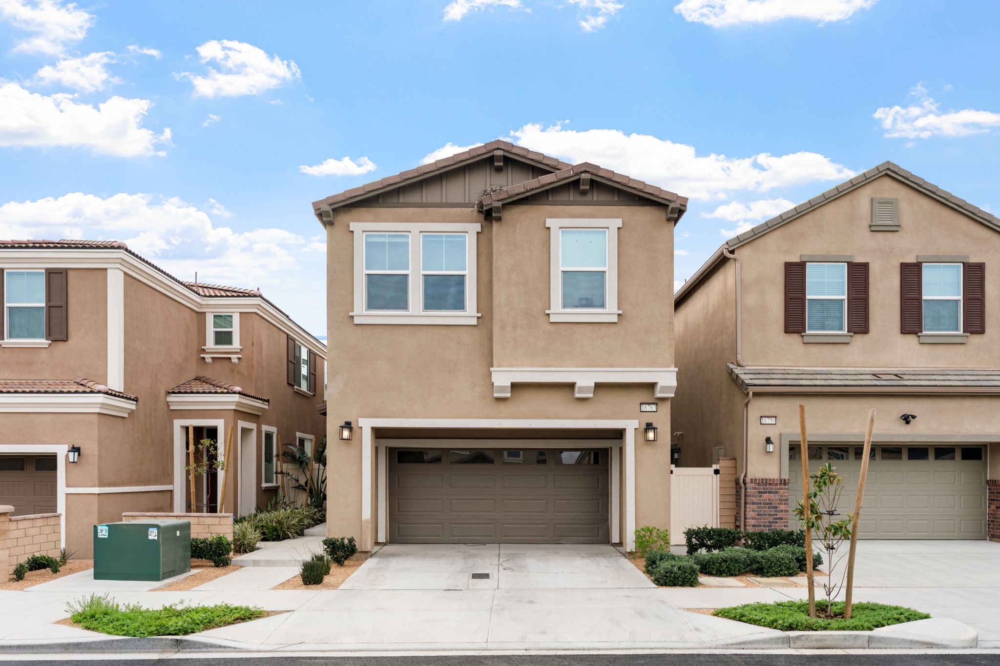 A modern two-story beige house with a garage, neighboring houses on both sides, and a clear blue sky with some clouds overhead.