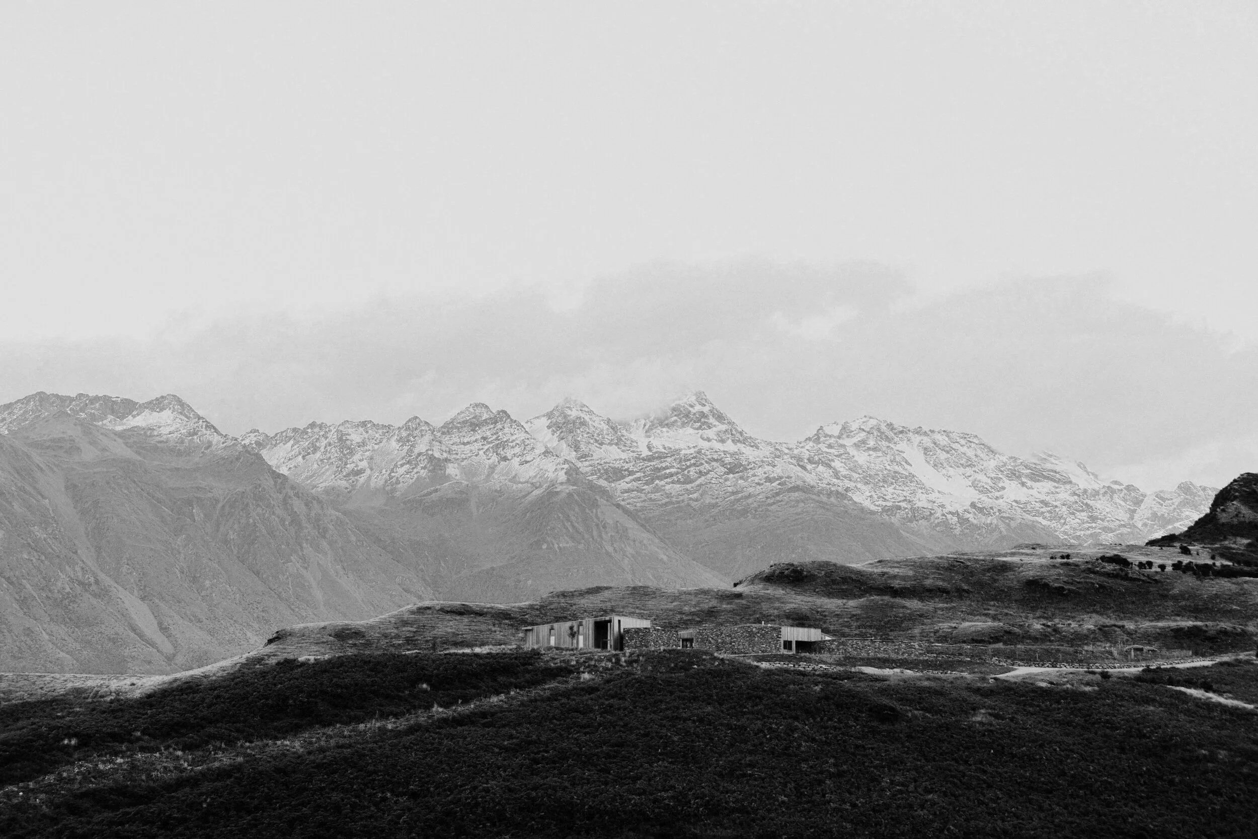 A black and white landscape photograph of snow-capped mountains in the background with a small estate in the foreground surrounded by grassy fields.