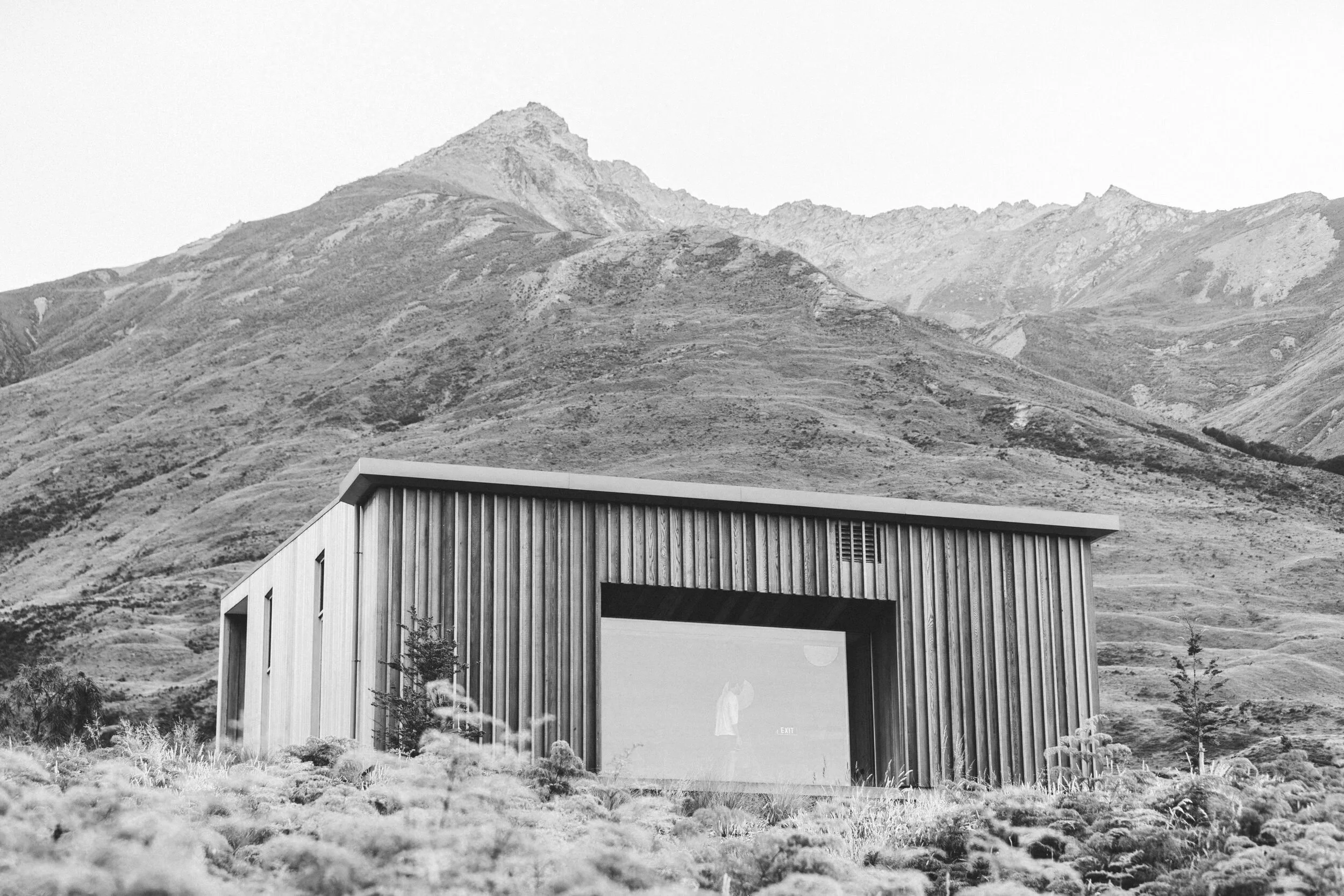A modern home with wooden vertical siding and a gabled roof, set against a backdrop of mountainous terrain with rocky mountain peaks.