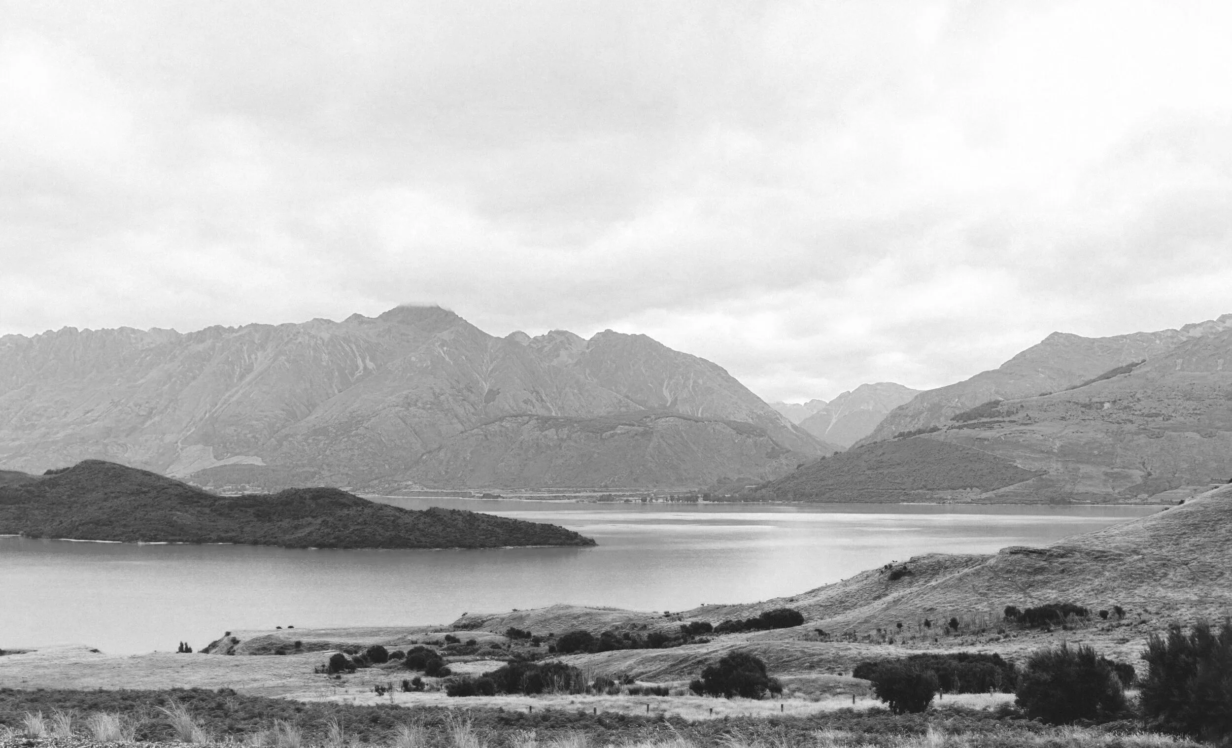 Scenic view of a lake property with mountains in the background, cloudy sky, and grassy hills in the foreground, captured in black and white.