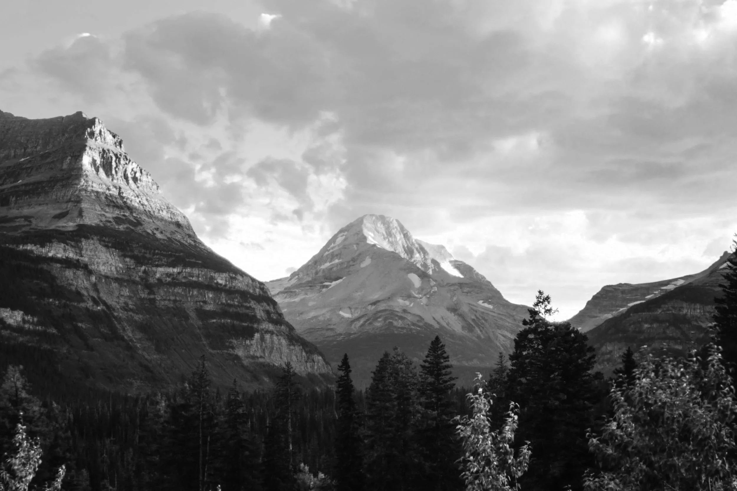 Black and white photograph of mountain peaks with rugged terrain and patches of snow, with a cloudy sky overhead and trees at the foreground.