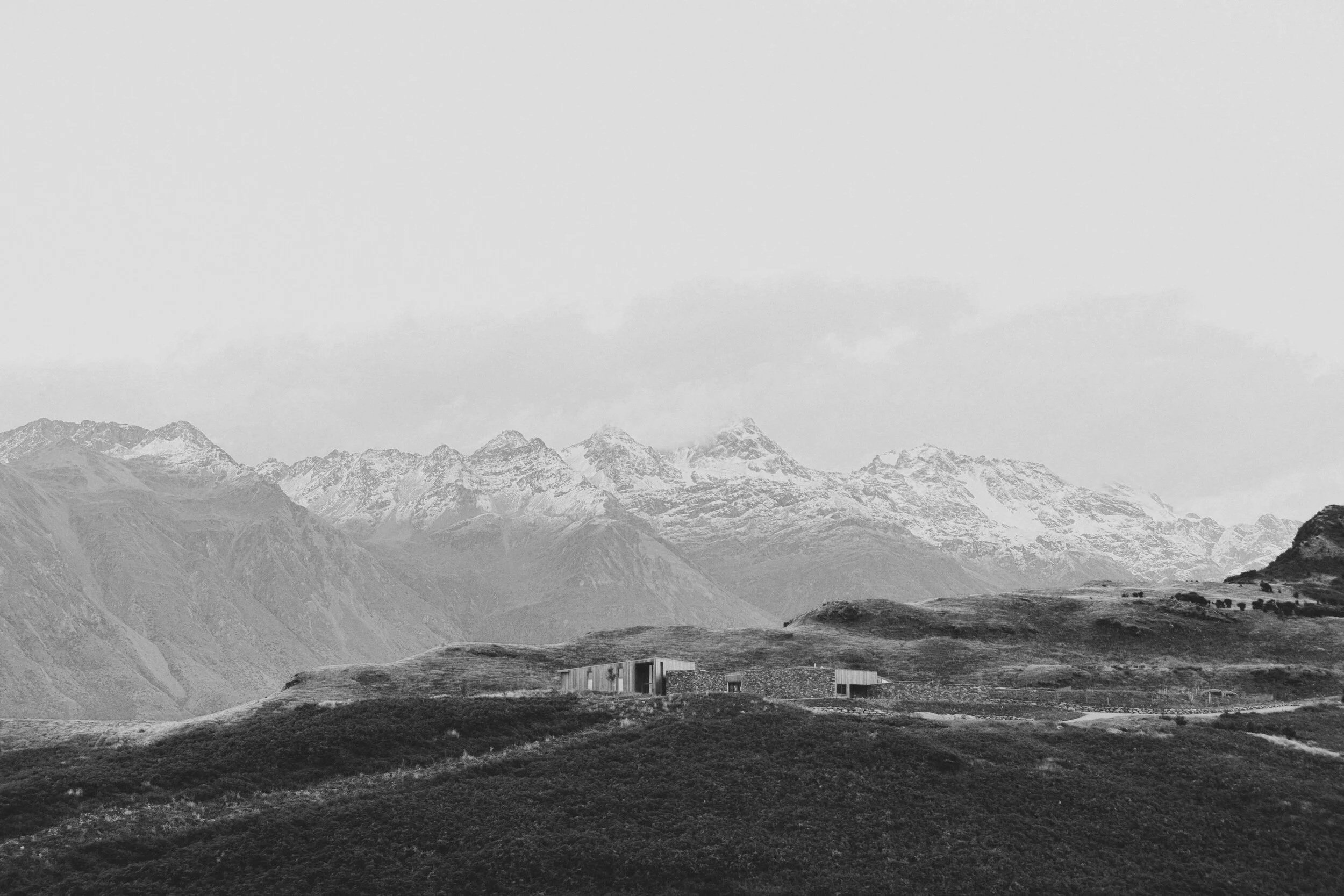 Black and white landscape photo of snow-capped mountains, rolling hills, and a small home in the foreground with a clear sky.