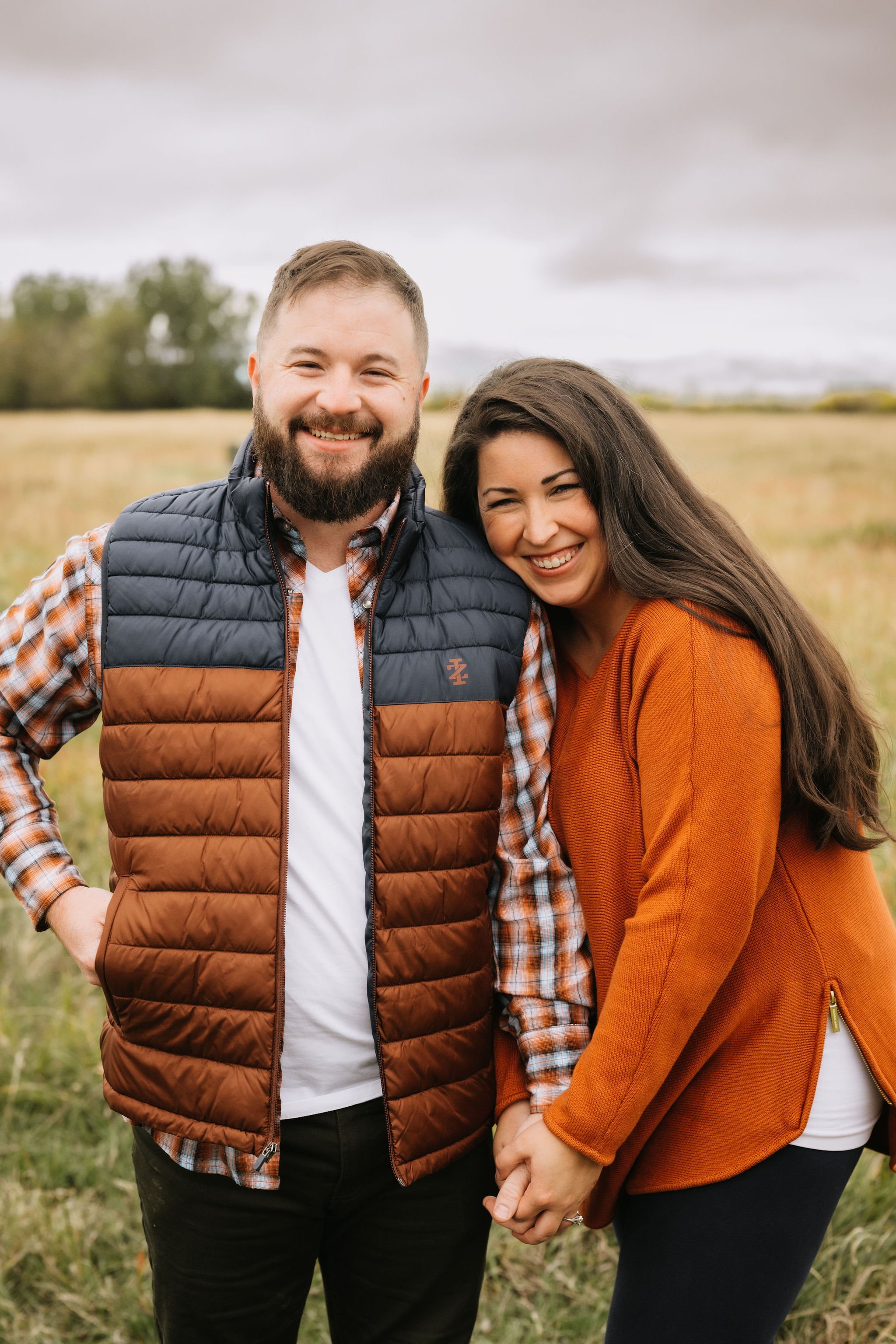 A smiling man and woman standing in Bozeman, MT, in a field with cloudy sky in the background, holding hands.