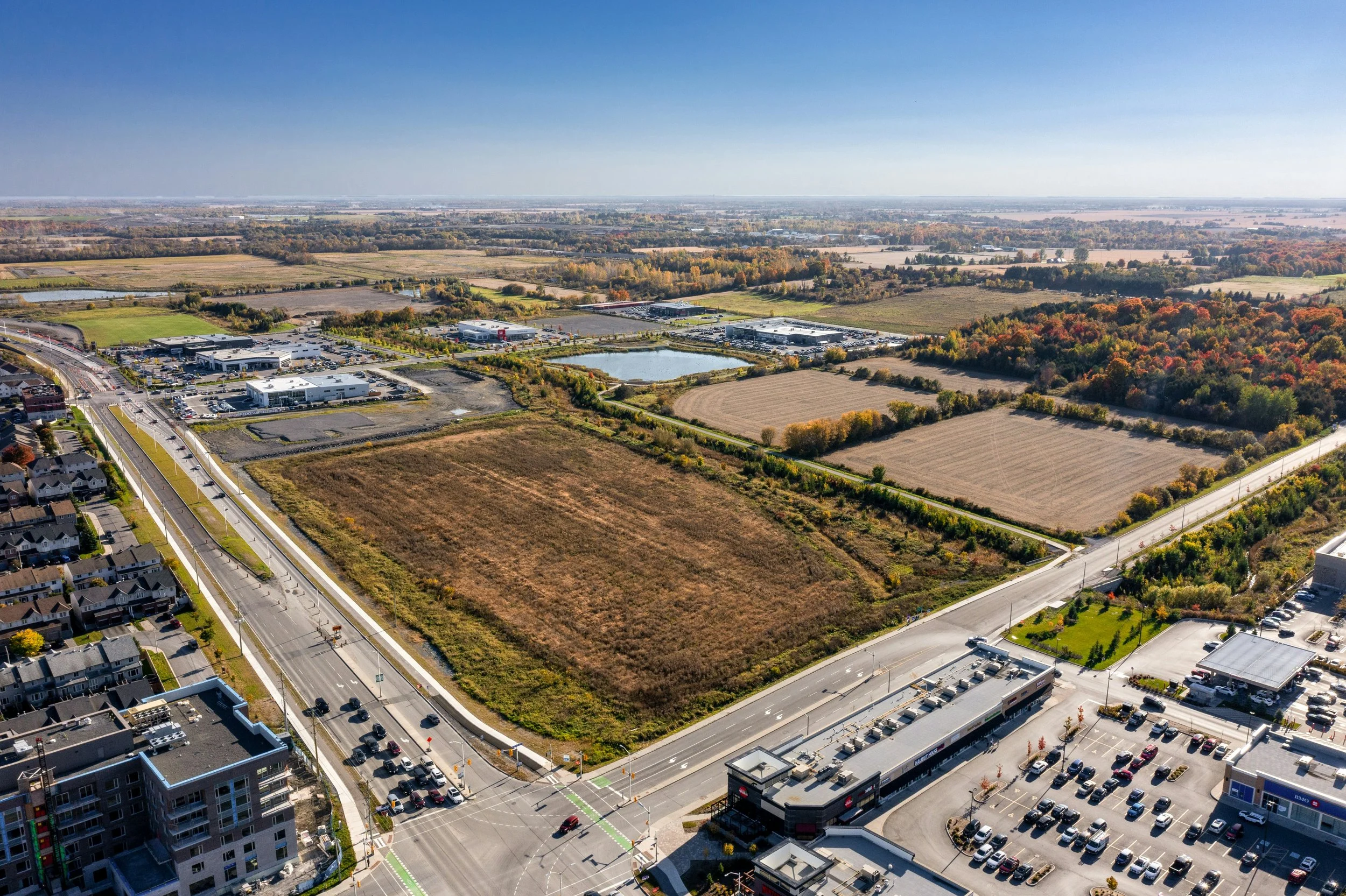Aerial view of a suburban neighborhood with a commercial shopping area, open fields, and a residential area amidst fall foliage.