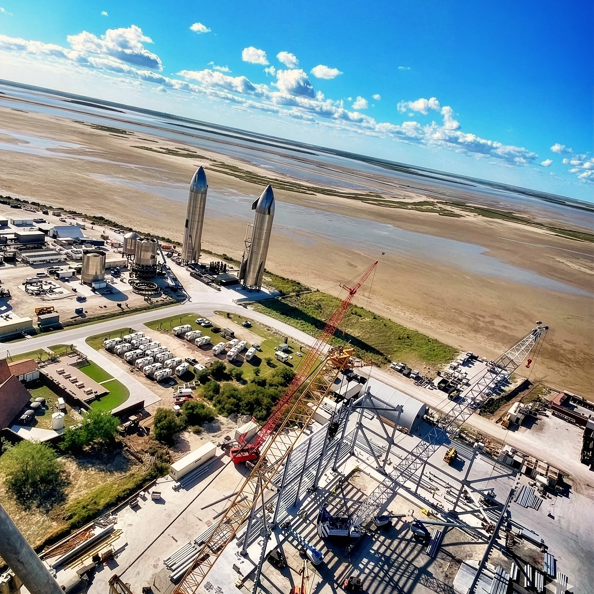 An aerial view of a coastal area with ships and construction site, featuring two large rockets, a crane, and parked vehicles, with wetlands and clouds in the background.
