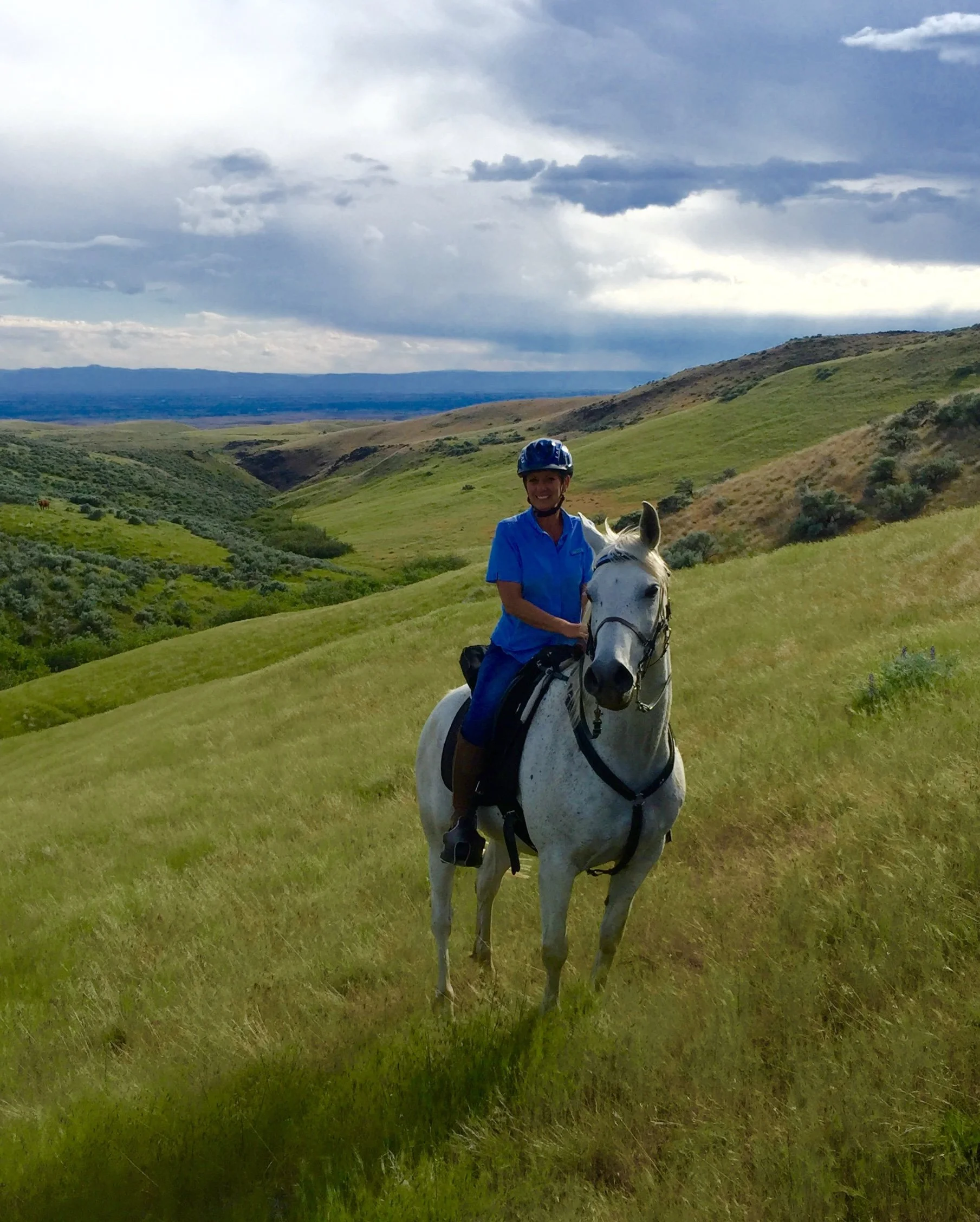 A person smiling while riding a white horse in a green hilly landscape with a cloudy sky in the background.