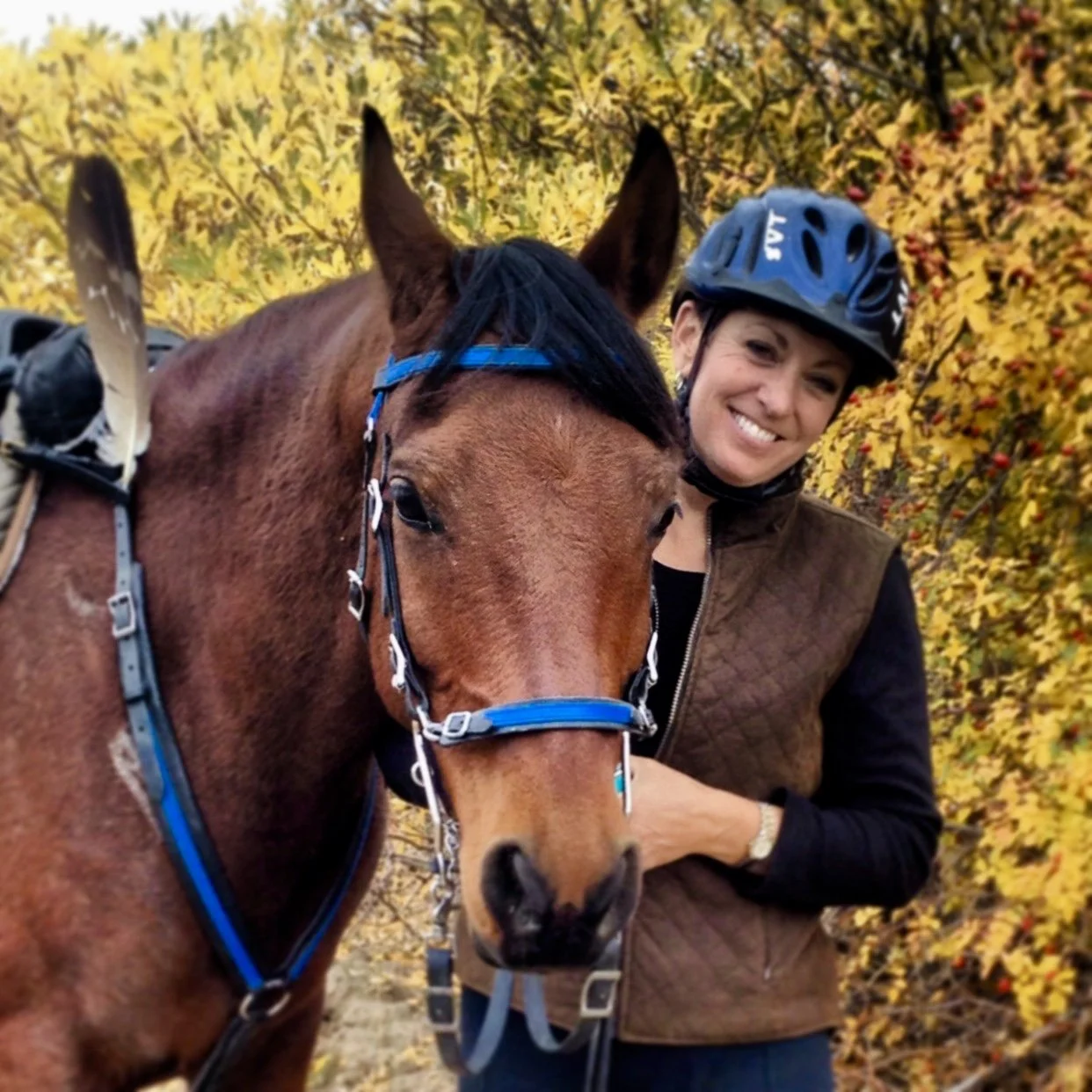 A woman smiling and wearing a helmet, standing next to a brown horse with a blue bridle, surrounded by yellow and orange autumn foliage.