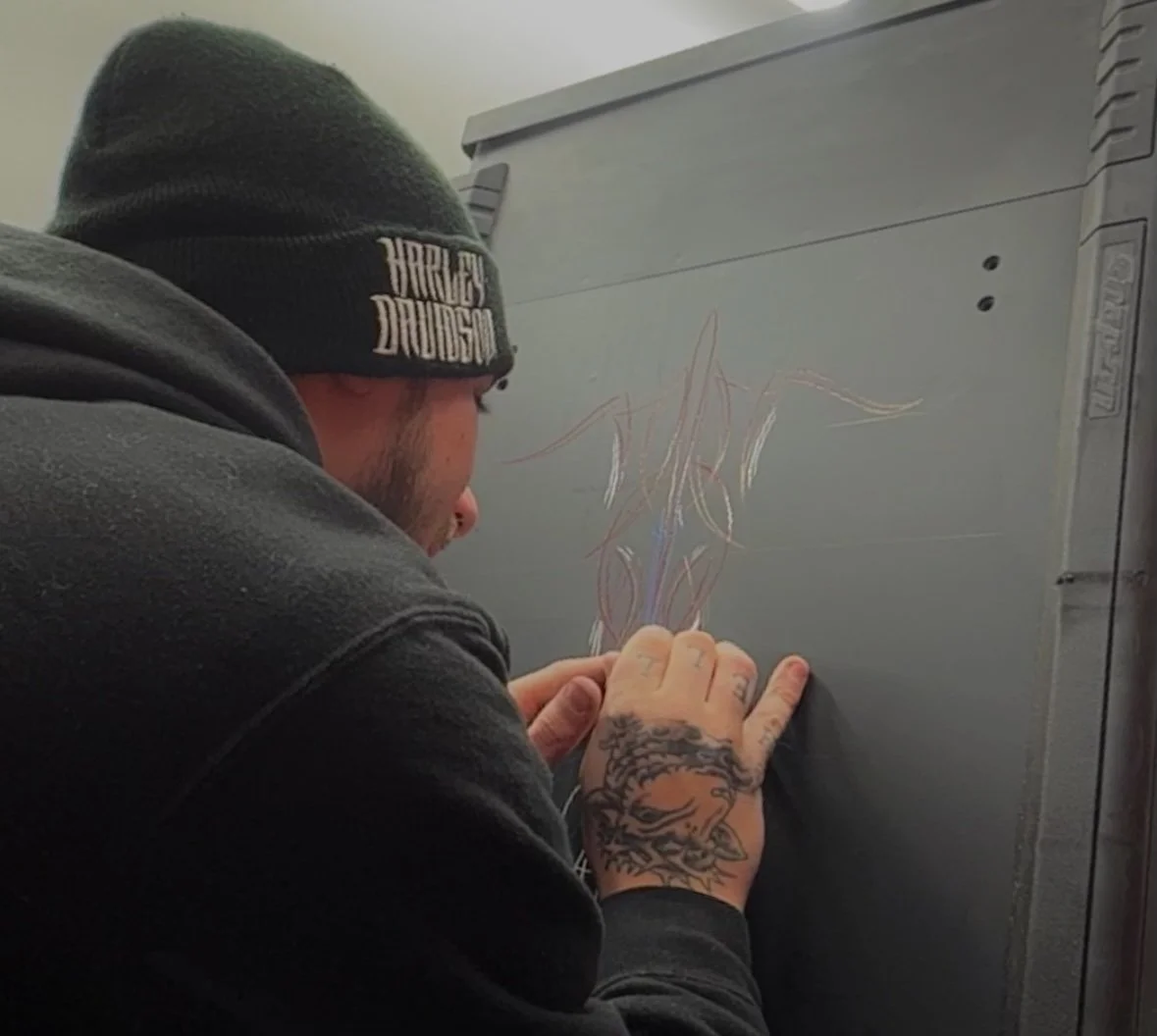 A man wearing a black beanie and black hoodie is drawing on a gray metal electrical box with colored chalk.