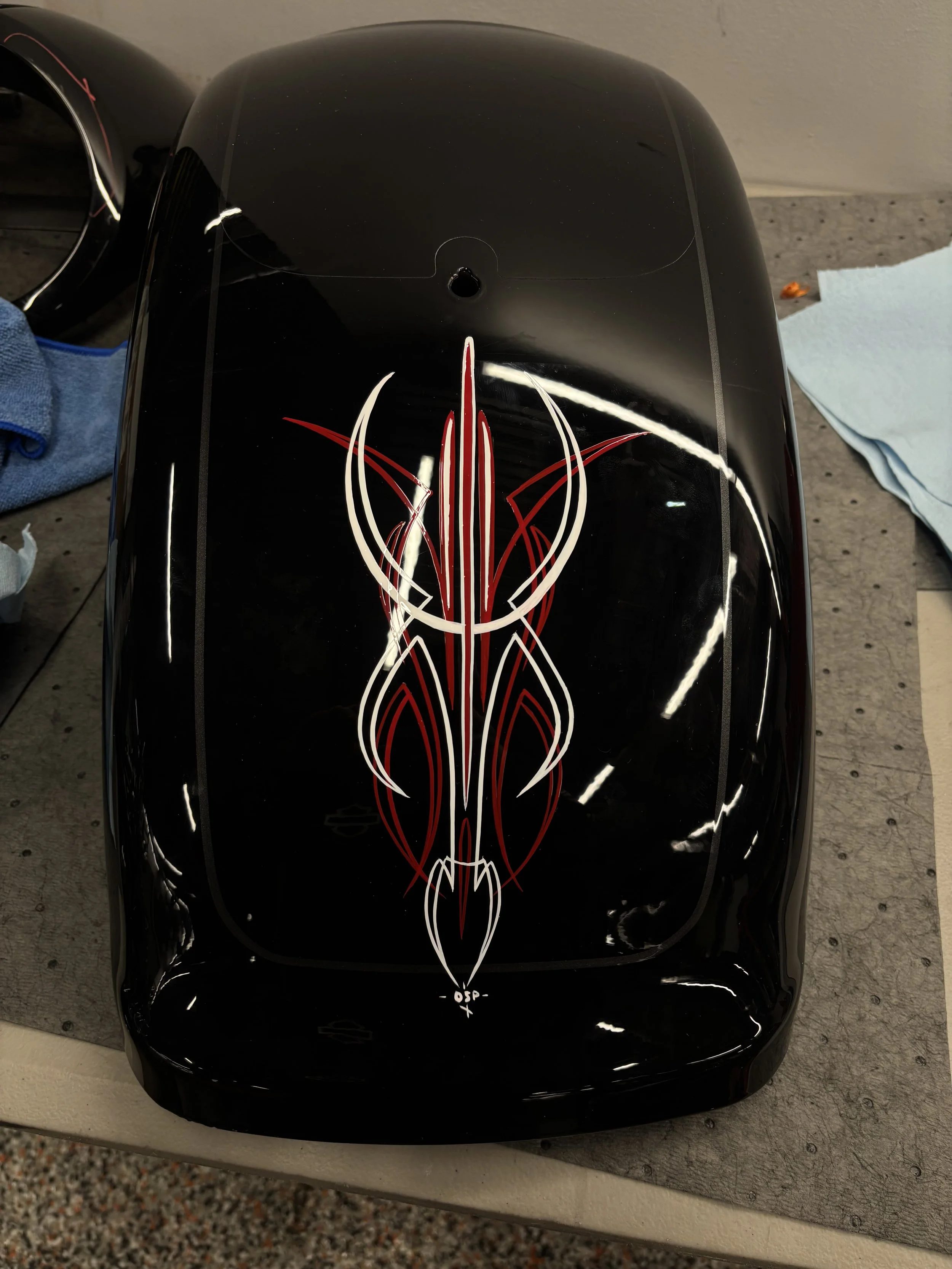 A black object, possibly a motorcycle fuel tank, with a red and white tribal-style decal design on it, placed on a workbench.