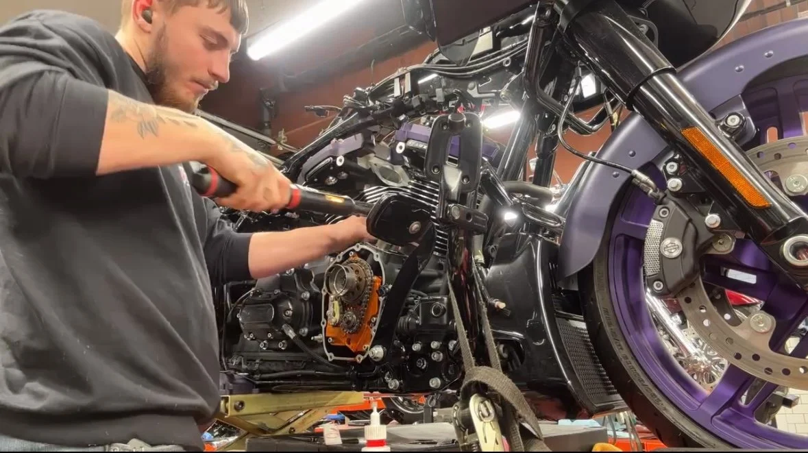 A man working on a black motorcycle's engine in a garage, with various tools and parts around him.