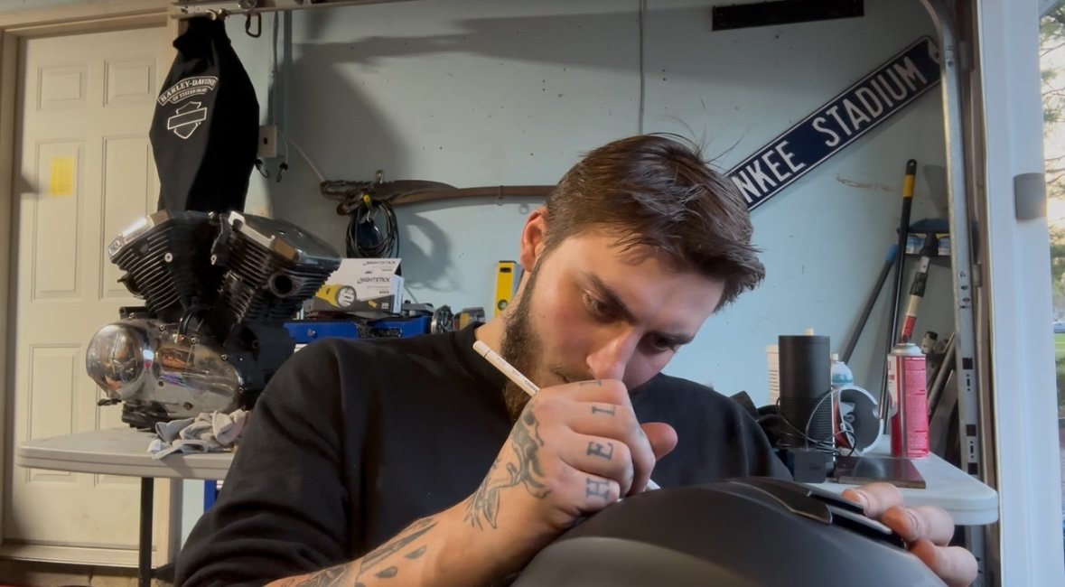 A man with tattoos and a beard writes on a motorcycle helmet with a white marker in a garage workshop. There is a large motorcycle engine in the background, along with various tools and equipment.