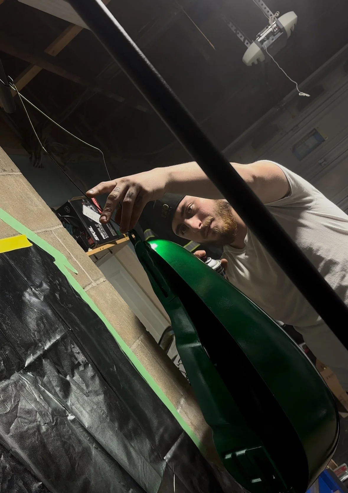 A man in a white t-shirt working on a green object, possibly a surfboard or similar item, in a workshop. The man is holding a tool near the object with a focused expression. The background includes shelves, a battery charger, and a garage door.