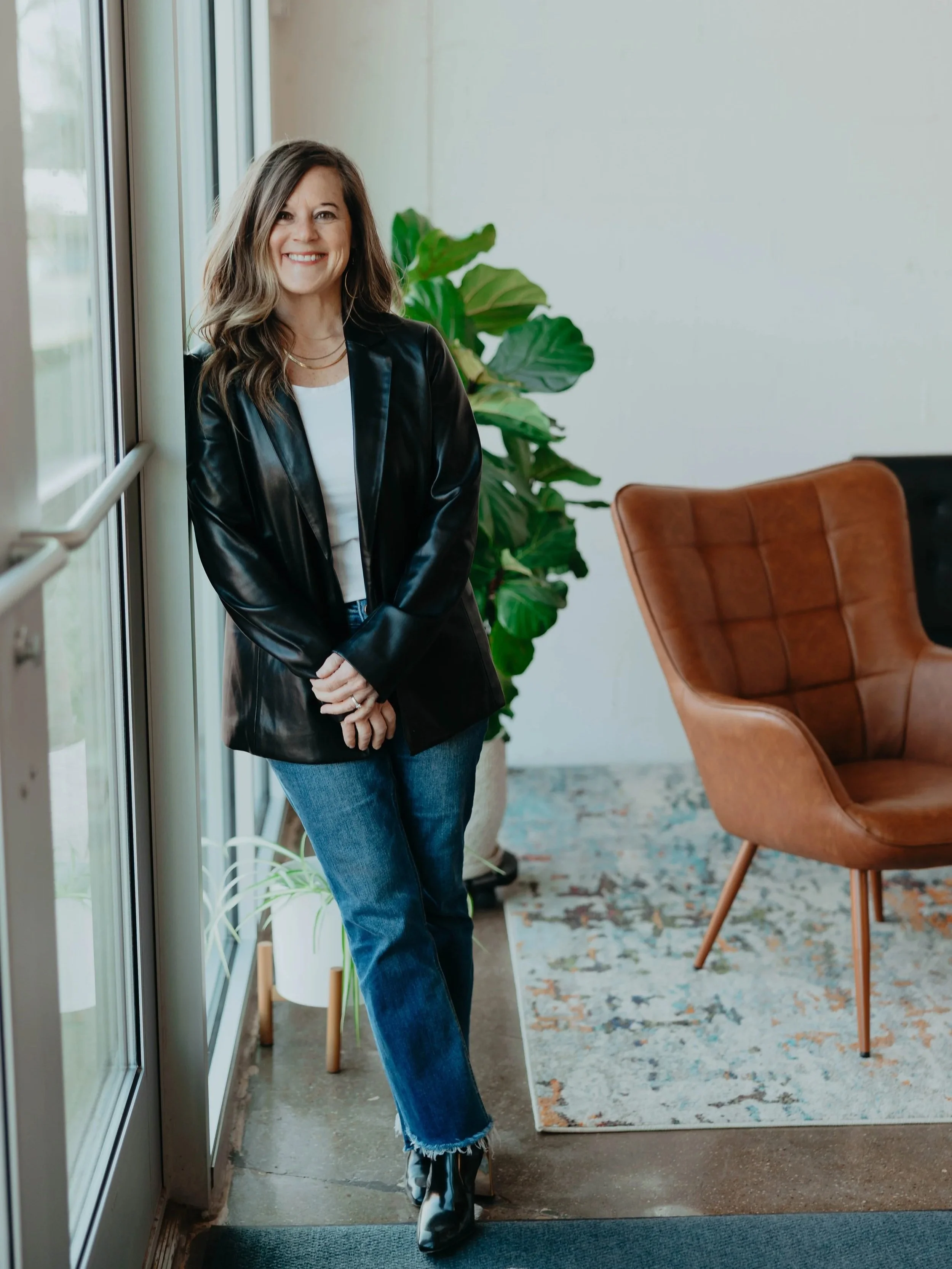 A woman with wavy brown hair smiling and standing near a large window in a modern interior with a brown leather chair, green plants, and a patterned rug.